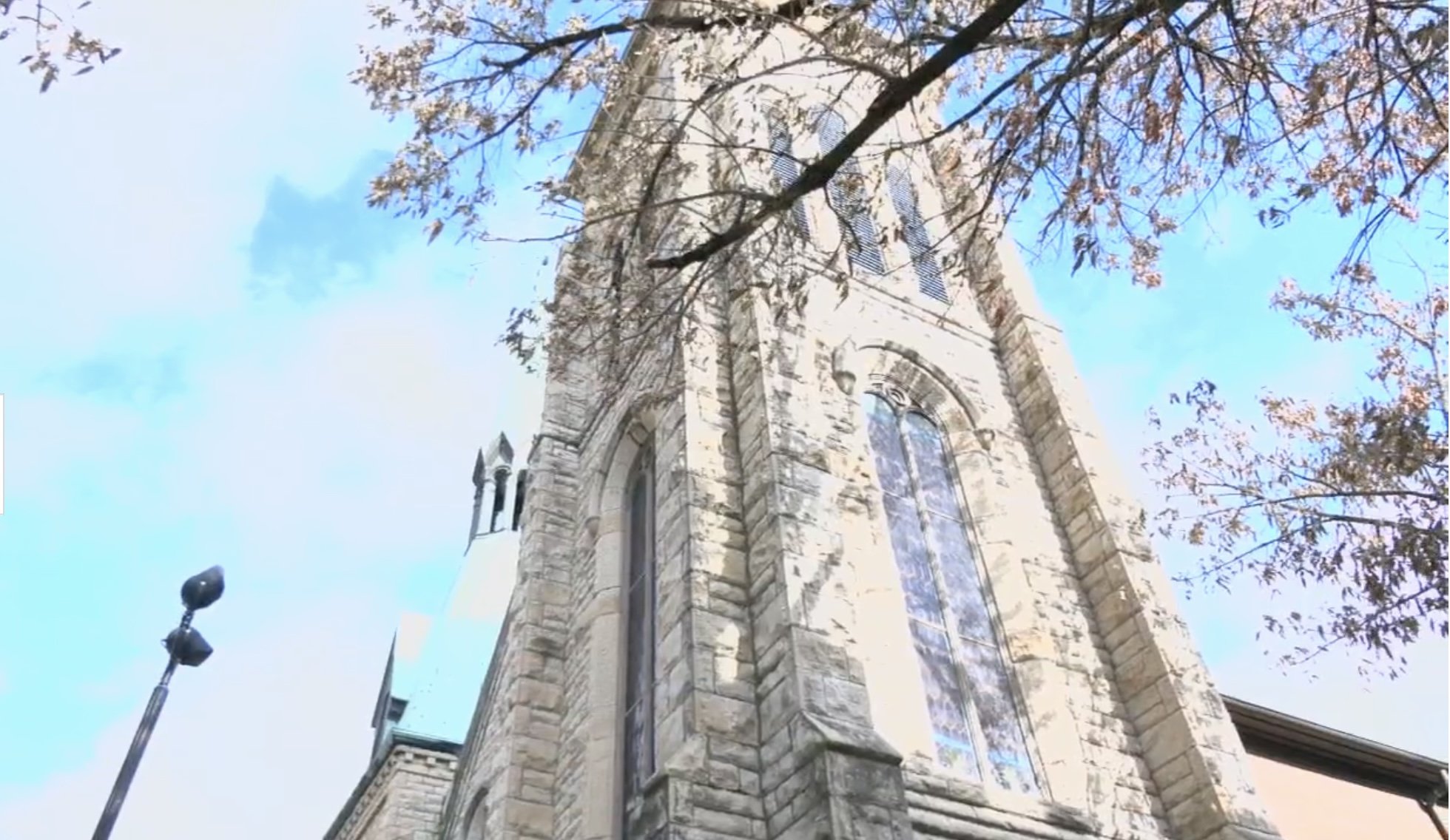 Low-angle view of a historic stone church steeple with tall arched windows and trees with autumn leaves in the foreground, against a bright blue sky with a few clouds.