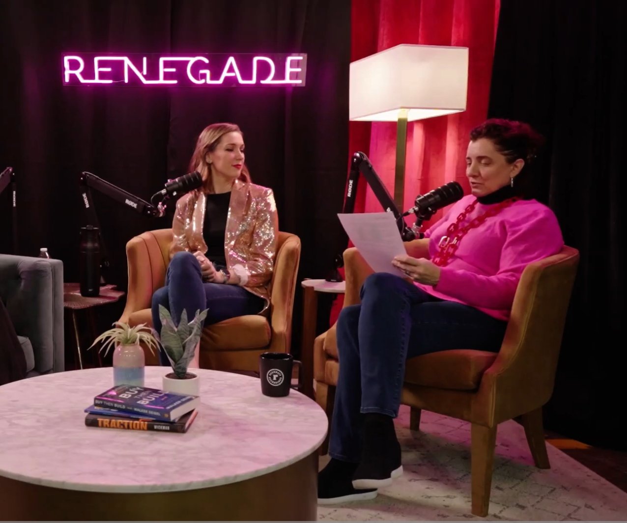 Two women seated in armchairs engaged in a podcast recording session. One woman is reading from a paper, while the other listens. Behind them is a neon sign that says "RENEGADE" and a large lamp. The setting includes a coffee table with books, a small plant, and a mug.