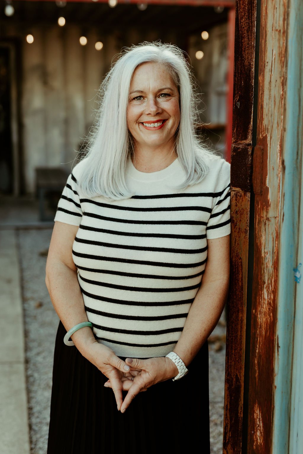 Smiling woman with long white hair wearing a white and black striped short-sleeve top standing outdoors beside a rusty doorframe.