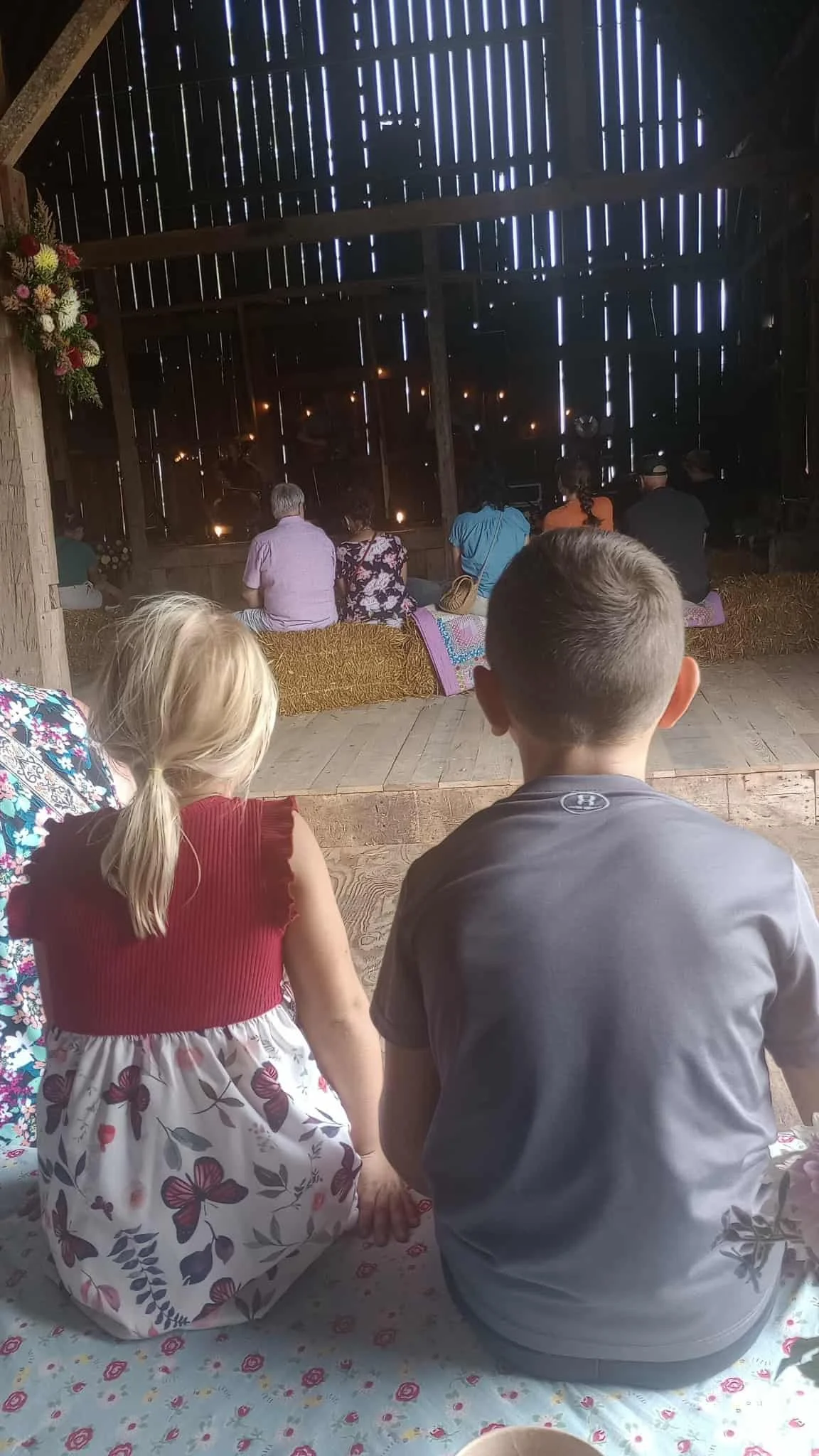 Two children, a girl with blonde hair and a boy with dark hair, are sitting and watching a puppet show in a barn with a rustic, wooden interior. The children are sitting on a blanket with floral patterns, and the barn's interior has a hay floor and wooden planks with gaps allowing light to shine through.