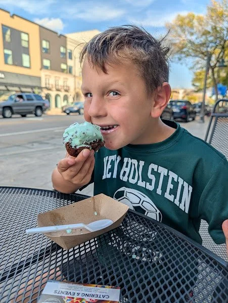 A young boy with short hair enjoying a chocolate and mint ice cream cone outdoors at a table, with a city street and buildings in the background on a sunny day.