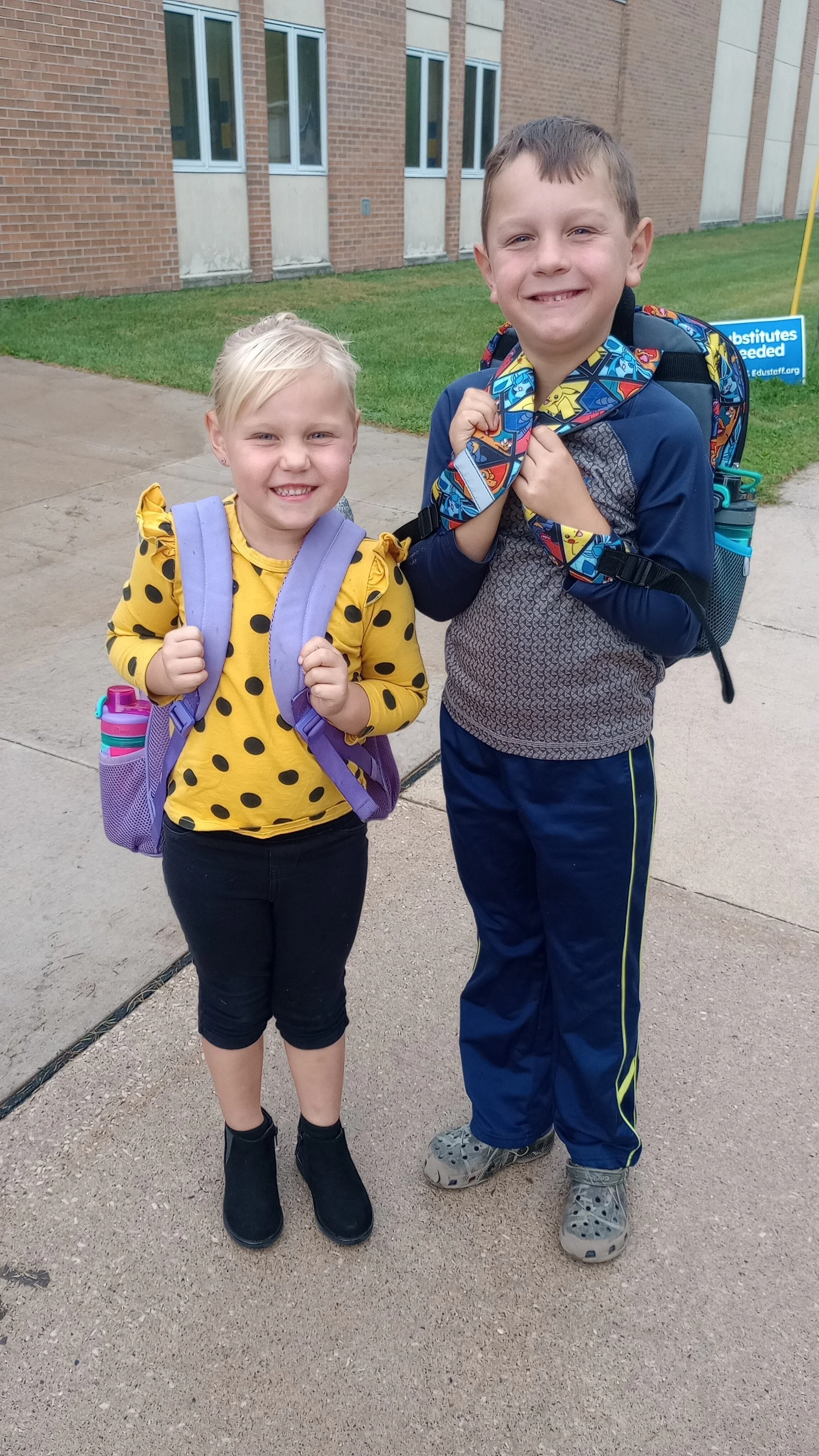 A young girl and a young boy standing outside a school building, smiling at the camera, each carrying backpacks.