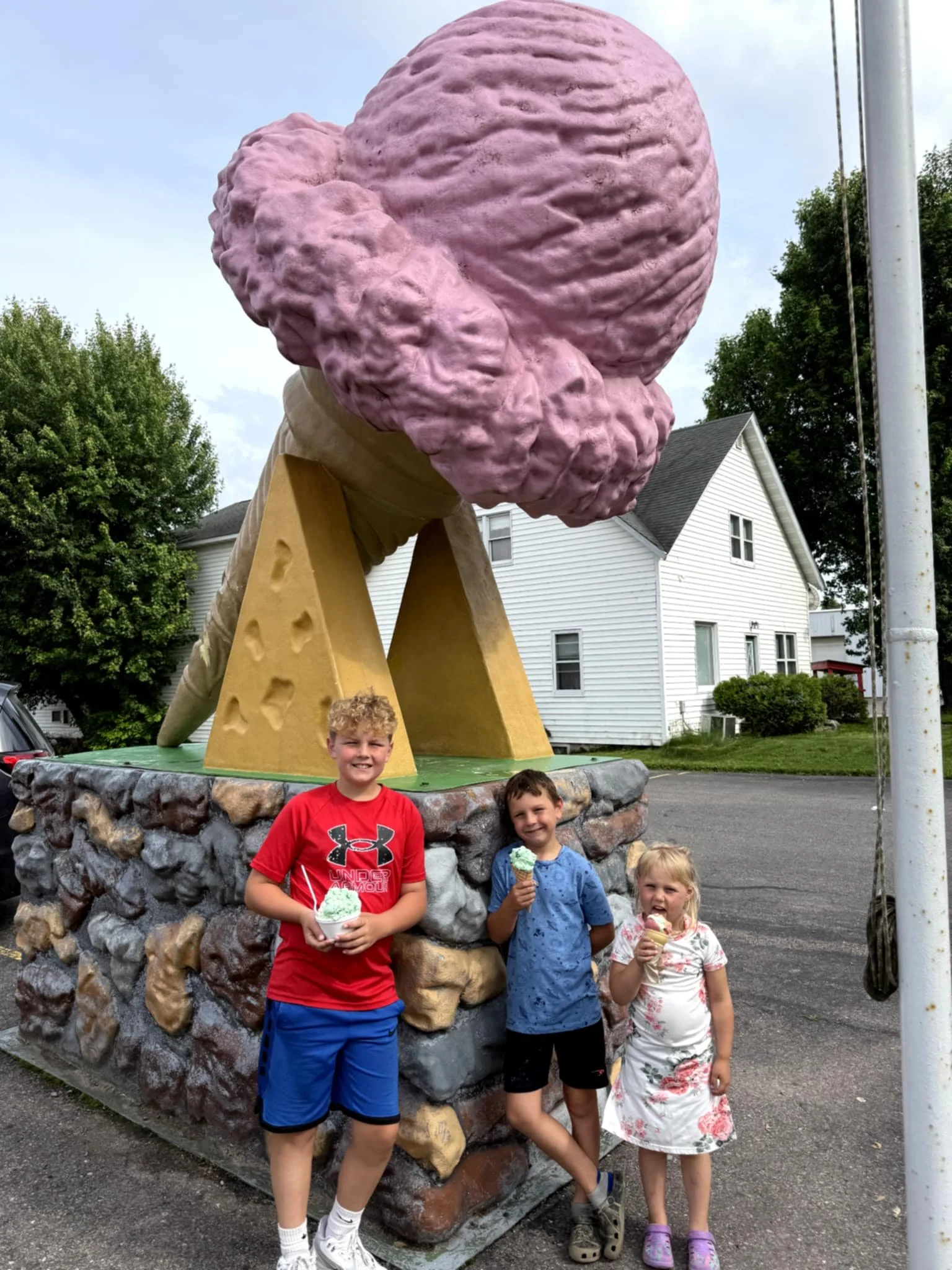 Three children standing in front of a giant ice cream cone sculpture, holding ice cream cones.