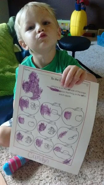 A young child with blonde hair and a green shirt holding a coloring worksheet showing multiple apple drawings, some filled in purple. The child is making a puckered face in a room with toys and a yellow toy in the background.