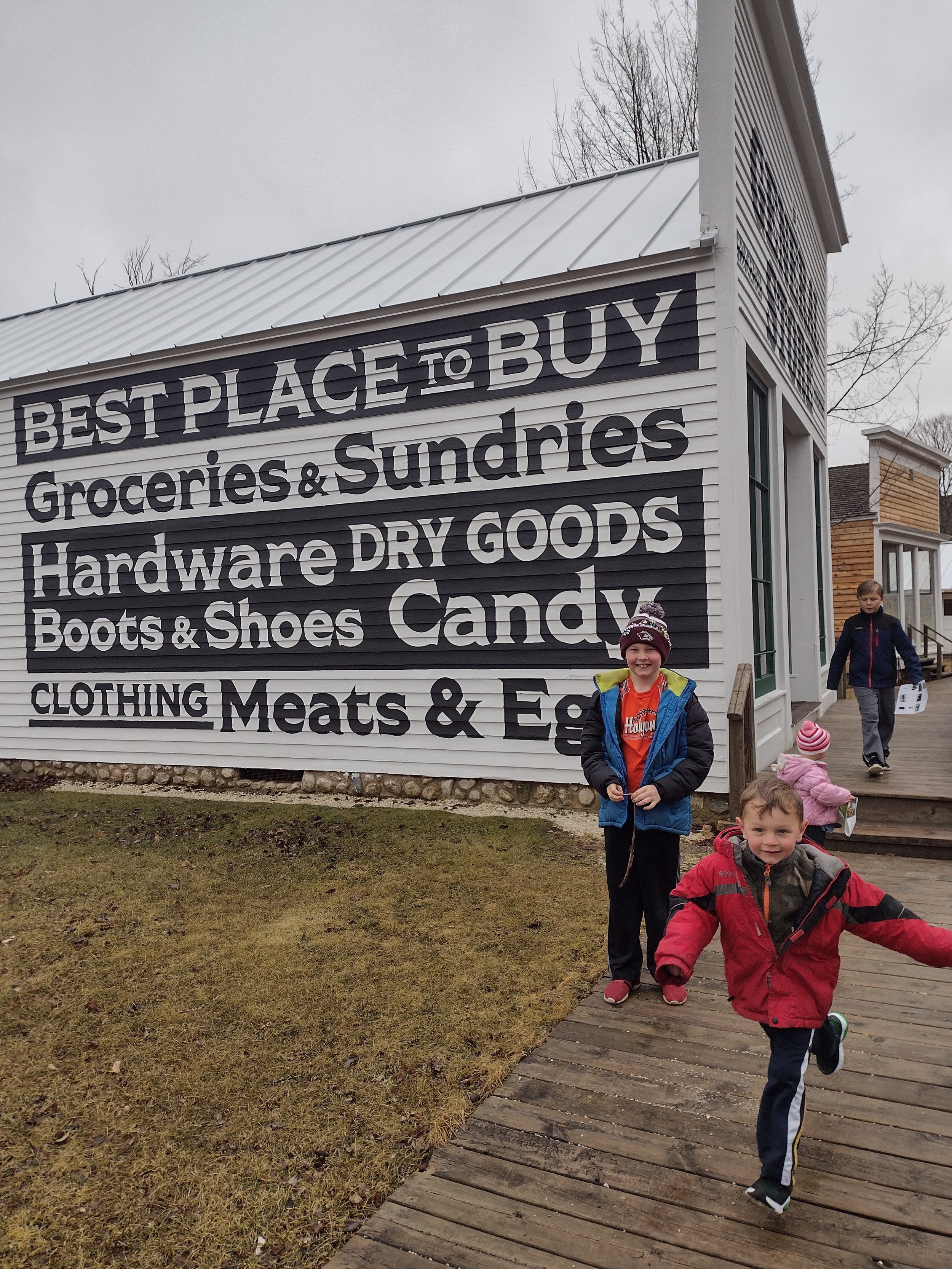 Children walking outside a store with a large sign that reads 'Best Place to Buy Groceries & Sundries, Hardware, Dry Goods, Boots & Shoes, Candy, Clothing, Meats & Eggs' on the building exterior.