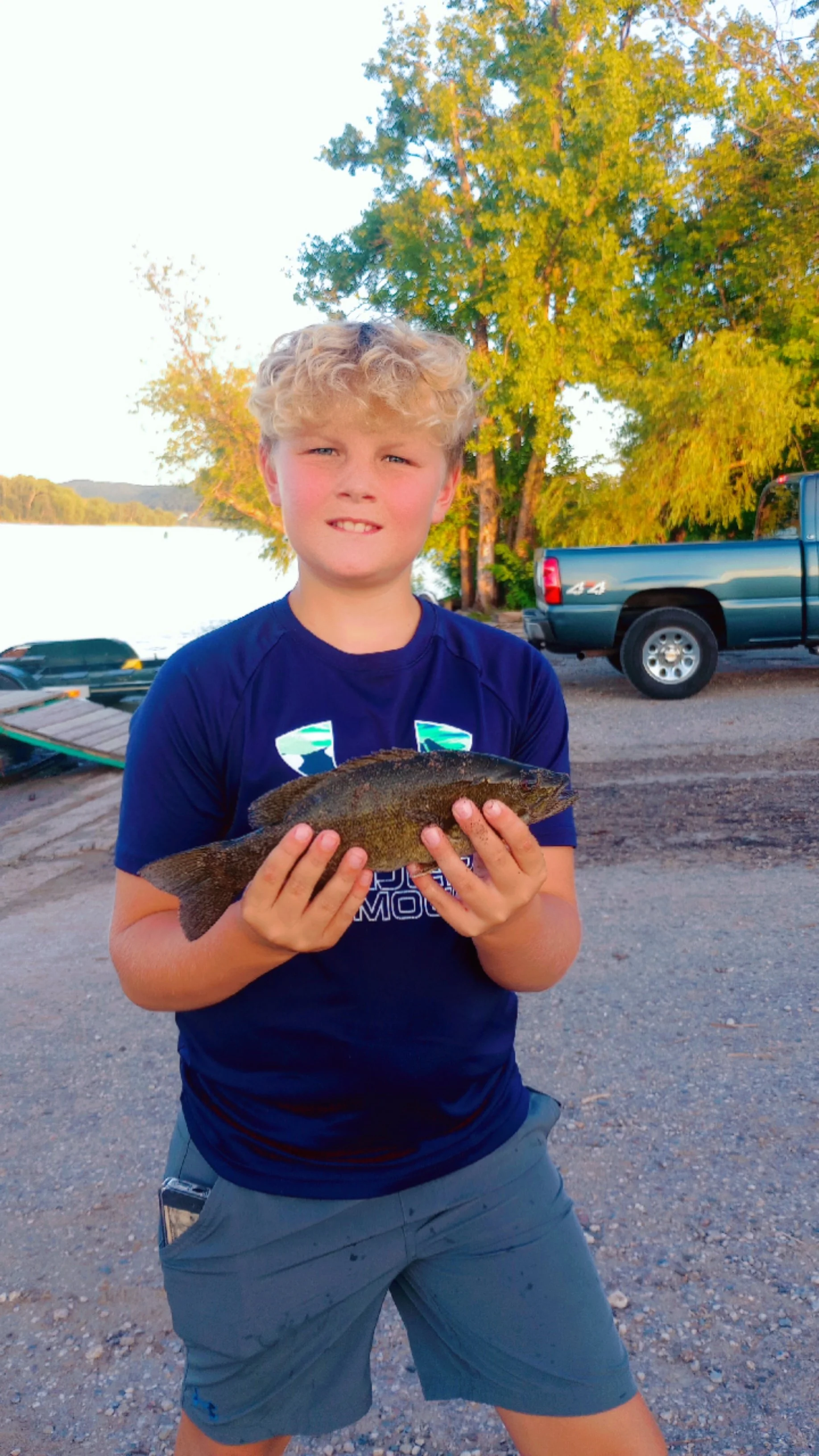 A young boy with blond curly hair holding a fish near a lake with trees and a blue pickup truck in the background.