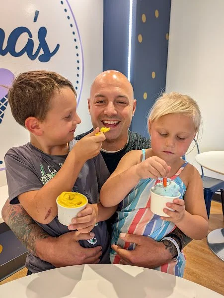 A man sitting with a young boy and girl, each holding cups of ice cream. The boy is licking his ice cream, and the girl is looking at her cup. They are indoors with a decorated wall in the background.