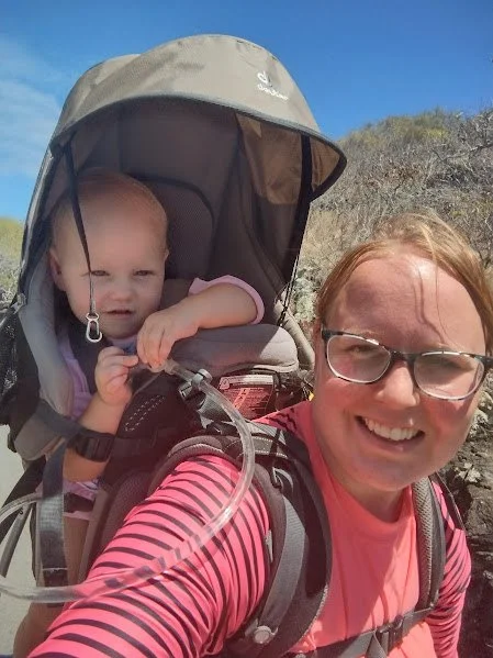Woman smiling while carrying a child in a child carrier on her back outdoors on a sunny day with a clear blue sky and rocky hill in the background.