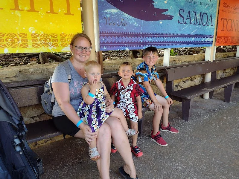 A woman and three children sitting on a bench in front of colorful signs about Samoa, with a stroller nearby.