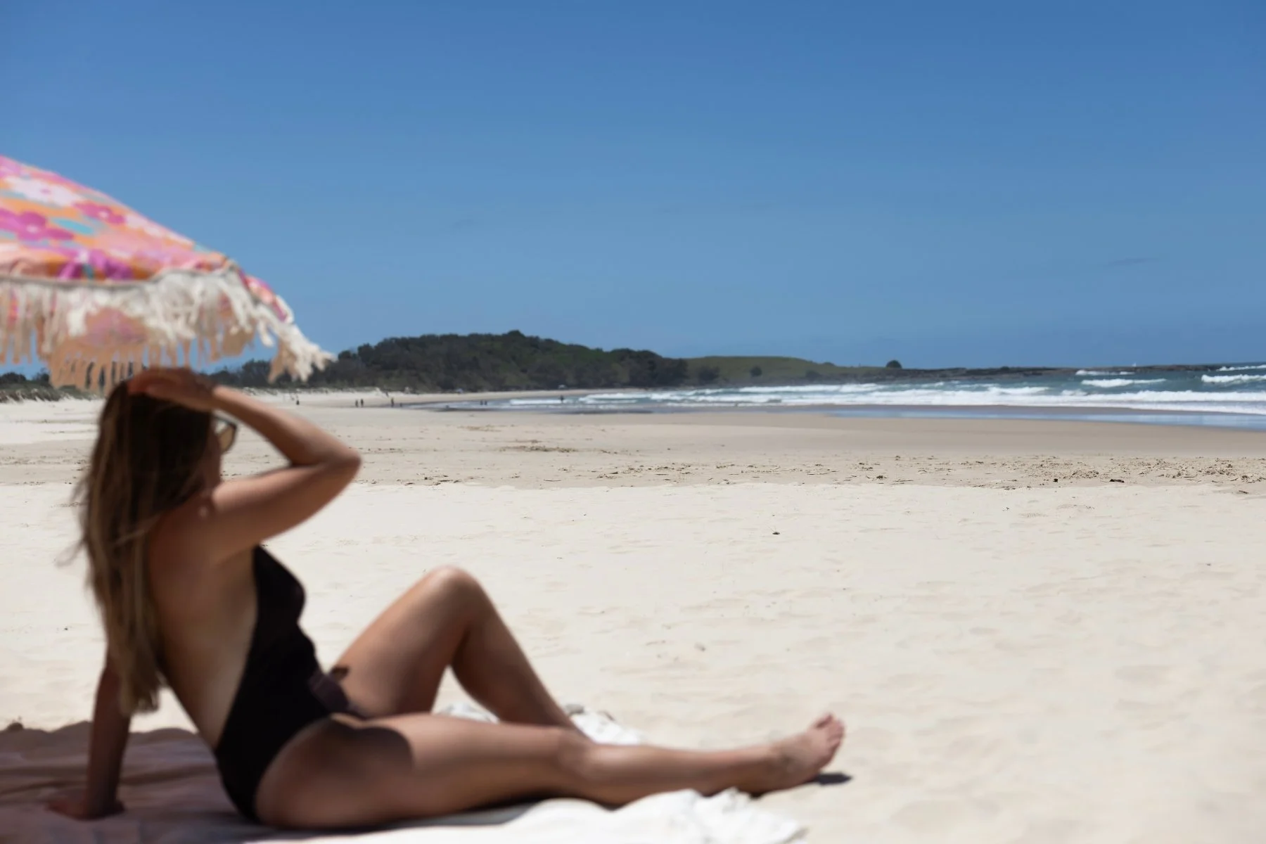 A woman in a brown swimsuit relaxes under a colorful umbrella on sandy Pippi beach, Yamba, gazing at the ocean waves under a clear blue sky.