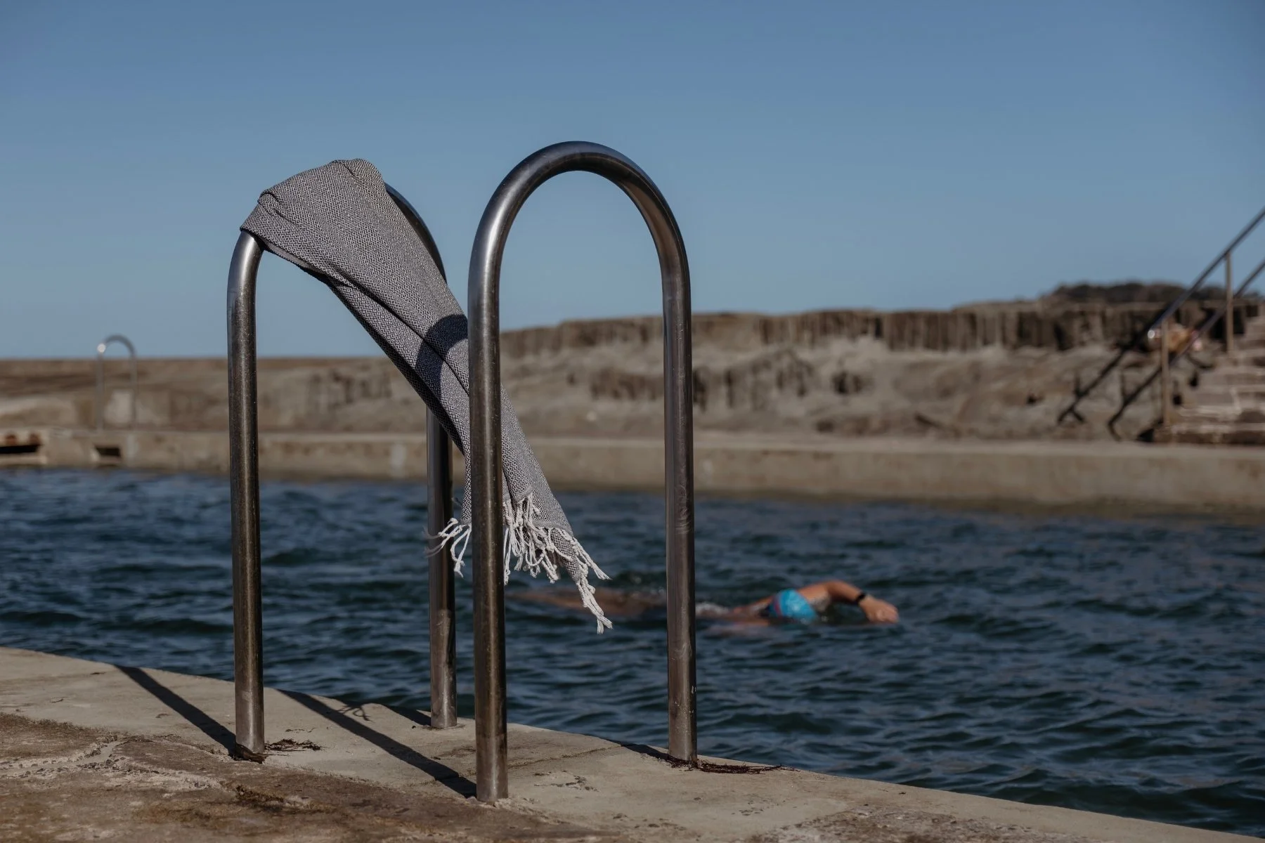 Swimming pool with metal ladder, draped beach towel, leading into the water. A swimmer is visible in blue swim cap in the pool, with rock shelf in the background.