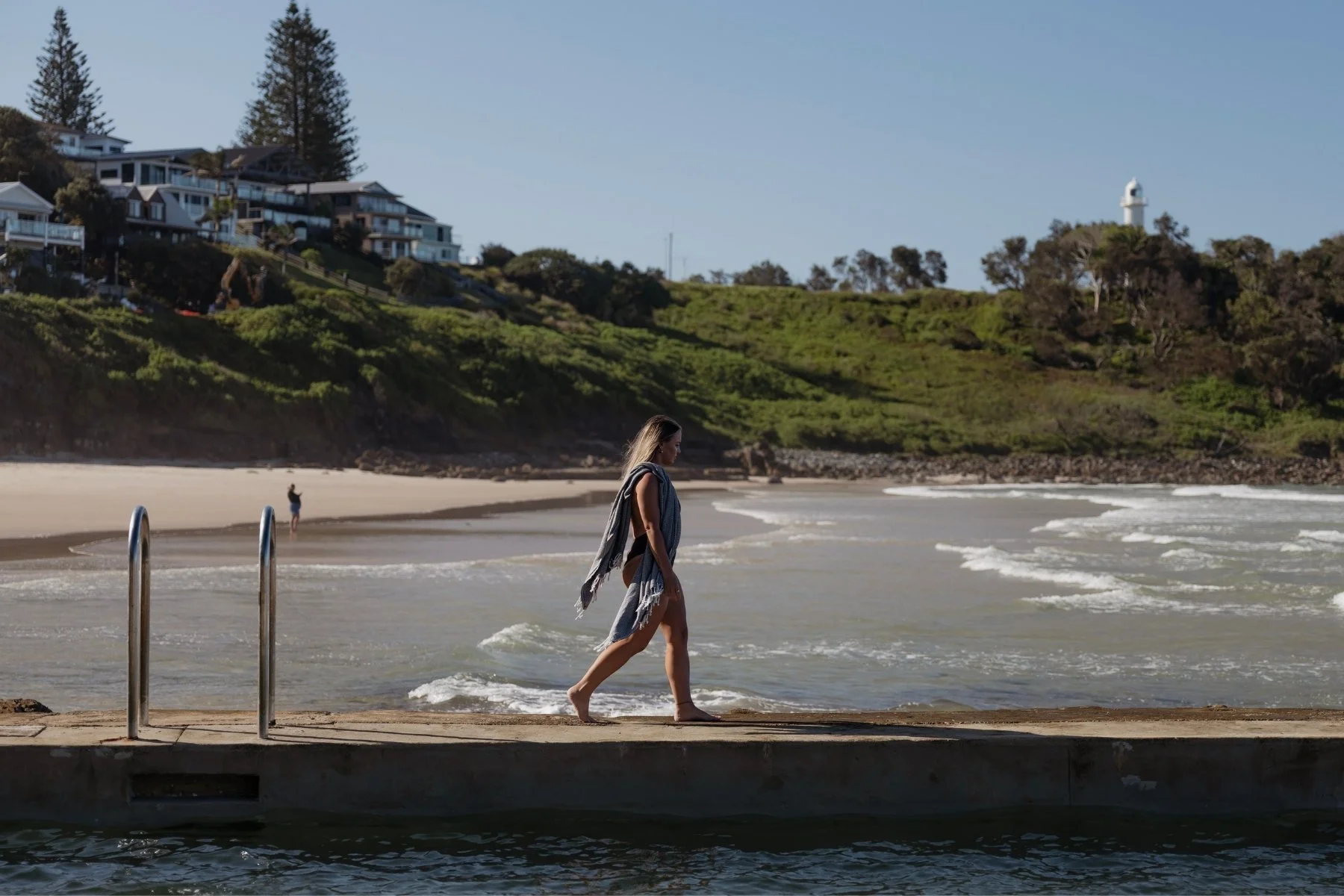 A woman walks barefoot along swimmng pool wall path by the beach, with waves gently lapping. In the background, lush greenery, houses, and a lighthouse dot the hillside.