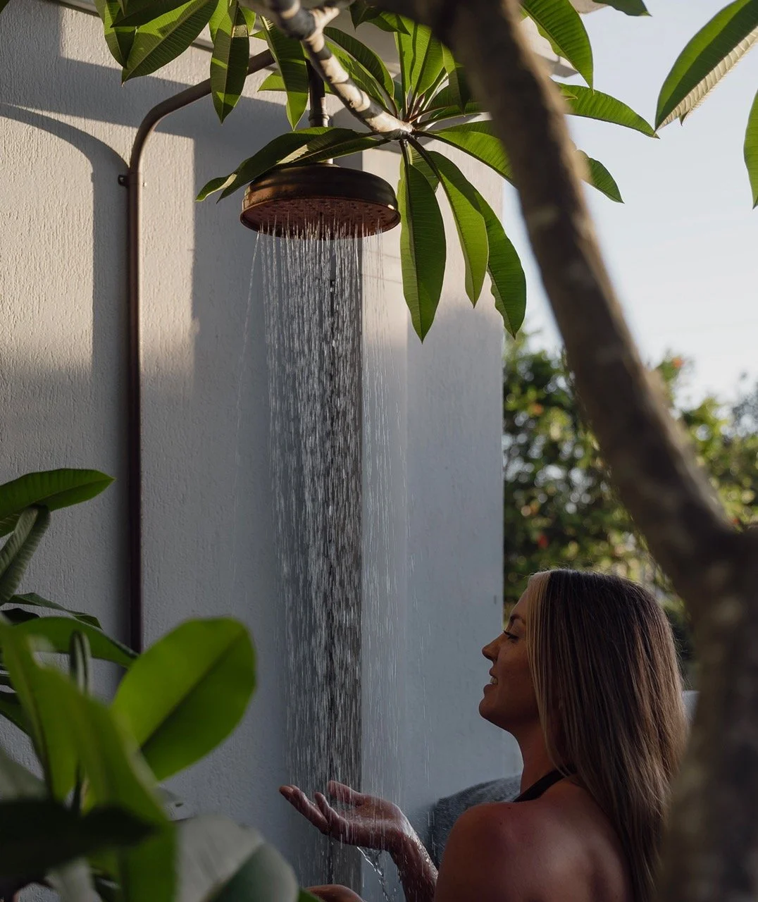 A woman enjoying an outdoor rainfall shower, with water cascading from a mounted copper showerhead surrounded by green plants.