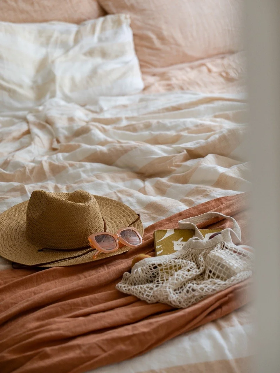 A bed with beige and white striped bedding, a beige wide-brim hat, pink sunglasses, a small box, a white mesh bag, and some personal belongings.