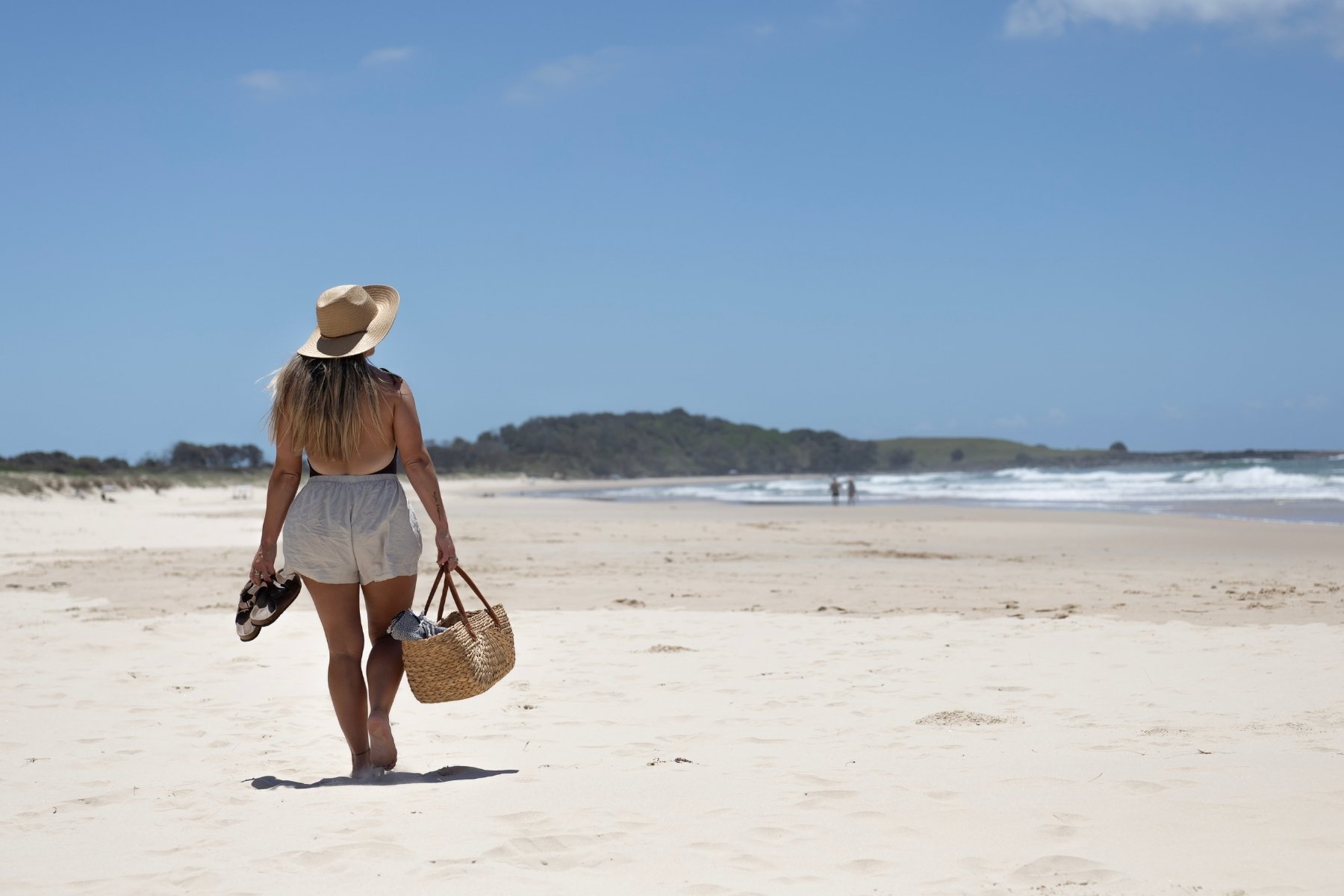 A woman in a sunhat walks along sunny Pippi beach Yamba, towards Lover's Point carrying a basket and sandals. The ocean is calm, and the scene feels tranquil and relaxed.