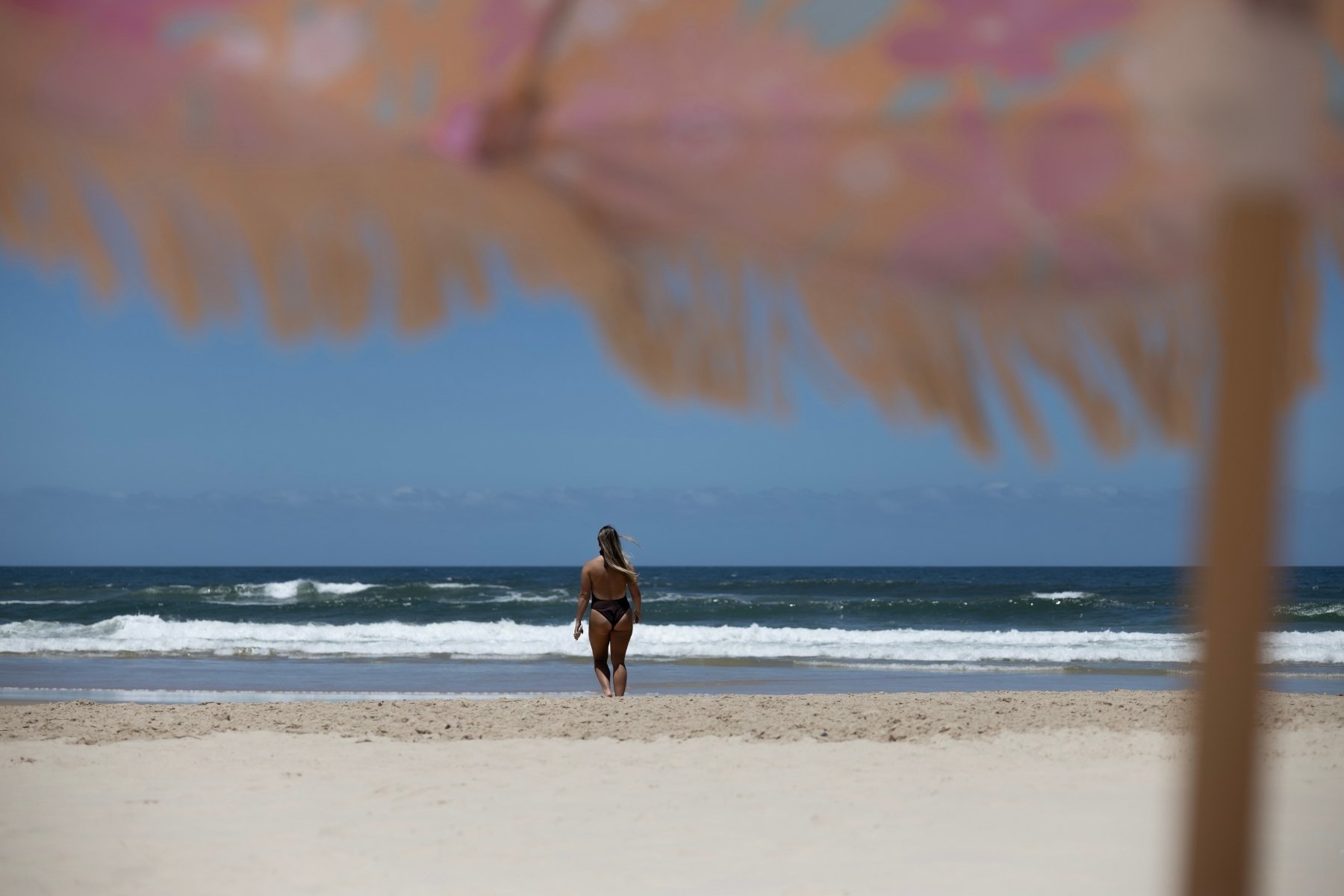 A woman in a swimsuit walks toward the ocean on a sandy beach. The scene is framed by a colorful, blurry beach umbrella, conveying a relaxed, sunny vibe.