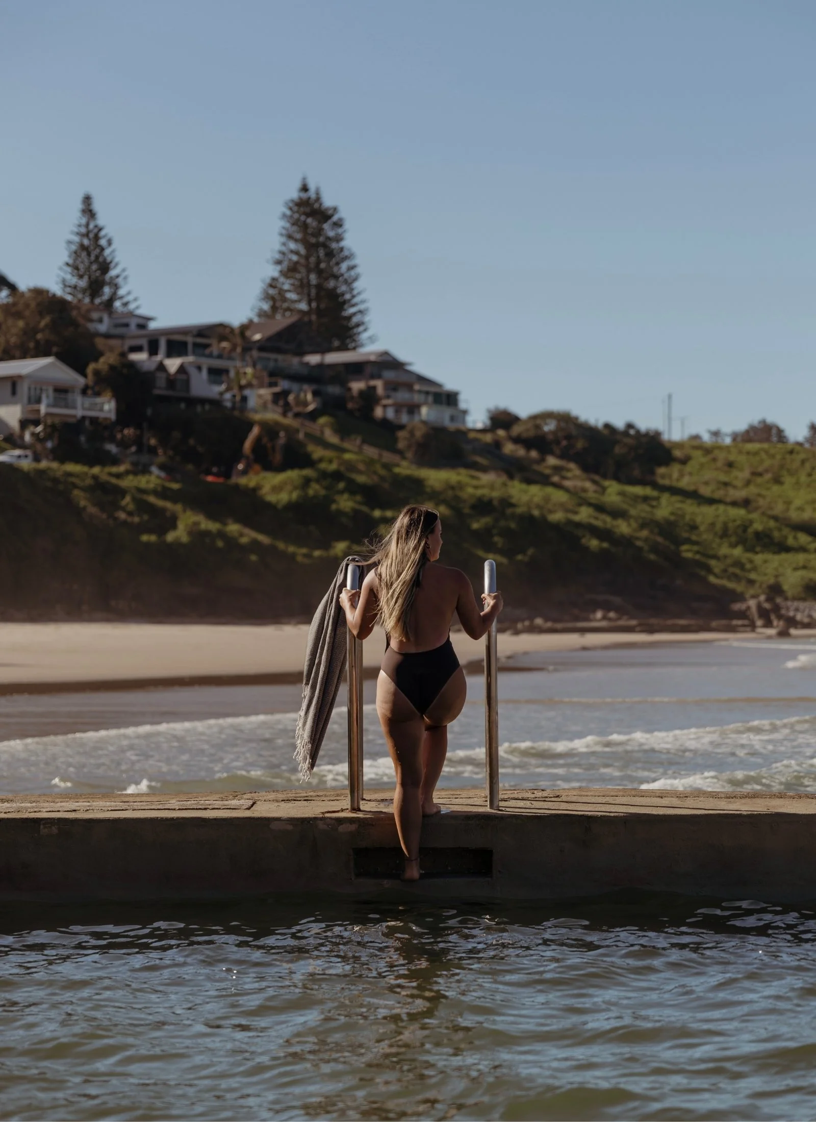 A woman in a brown swimsuit stands at the edge of main beach pool in Yamba, holding a towel. She faces a sunny beach with houses and greenery in the background.
