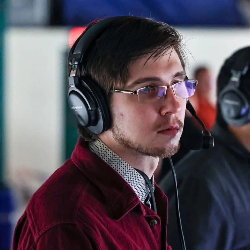 Young man with glasses and facial hair wearing a headset with a microphone, sitting in a busy environment.