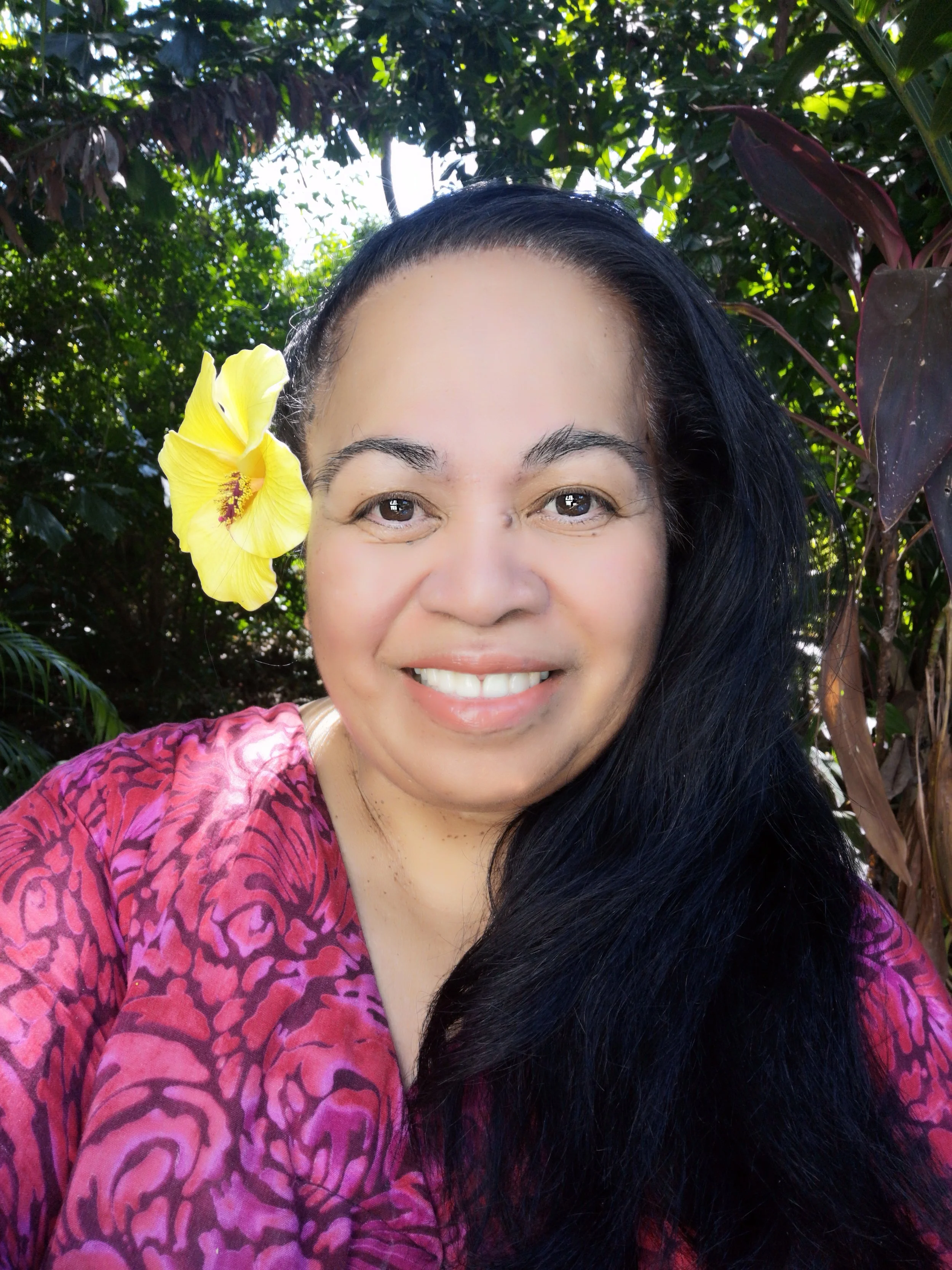 A woman smiling outdoors with a yellow hibiscus flower in her hair, wearing a pink patterned top, surrounded by green foliage.