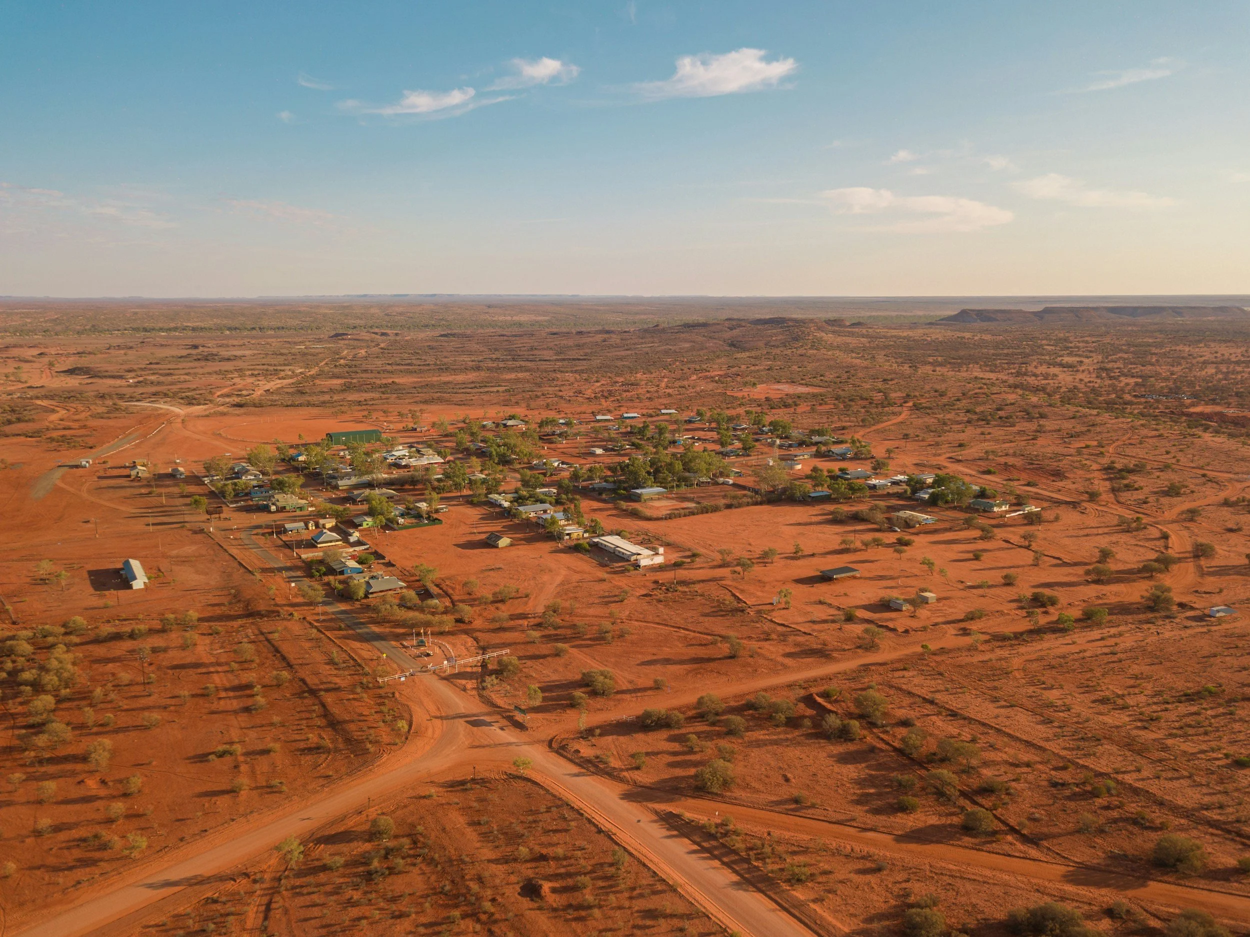 Aerial view of a small rural town in a desert landscape with red soil, sparse trees, and simple buildings, under a clear blue sky.