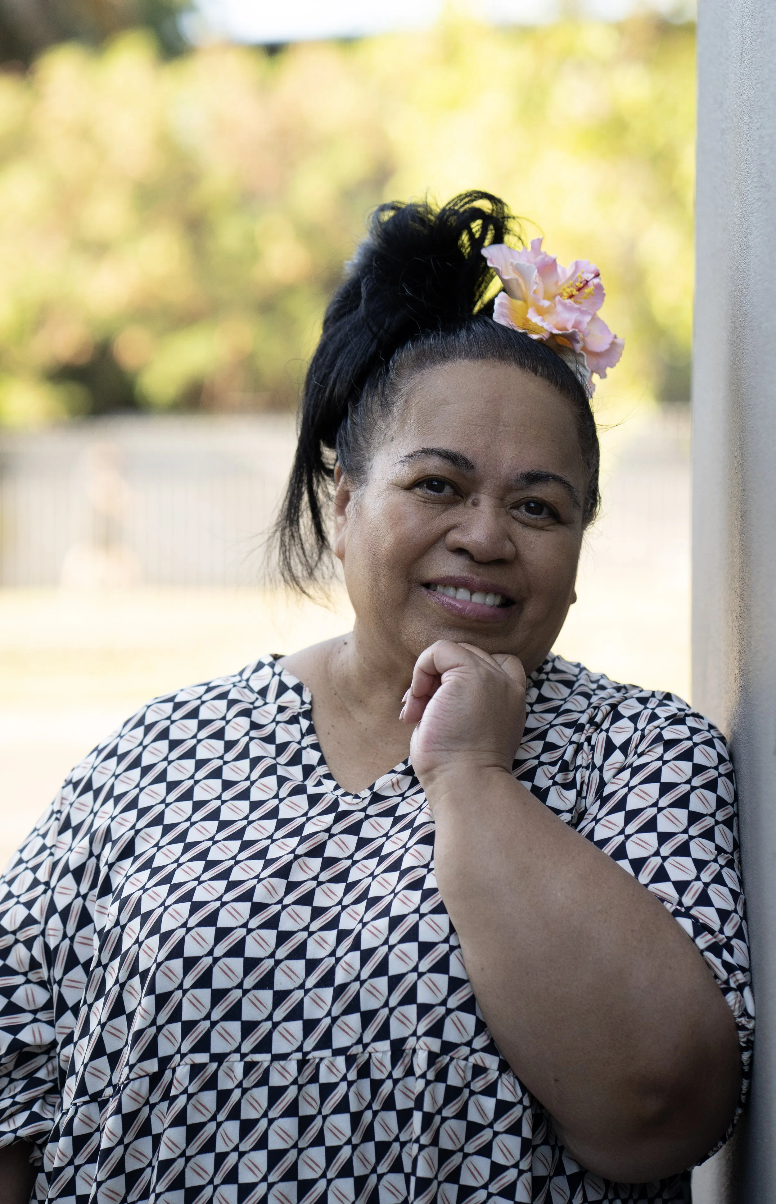 Smiling woman with dark hair styled up, decorated with pink flowers, wearing a black and white geometric patterned top, leaning against a wall outdoors.
