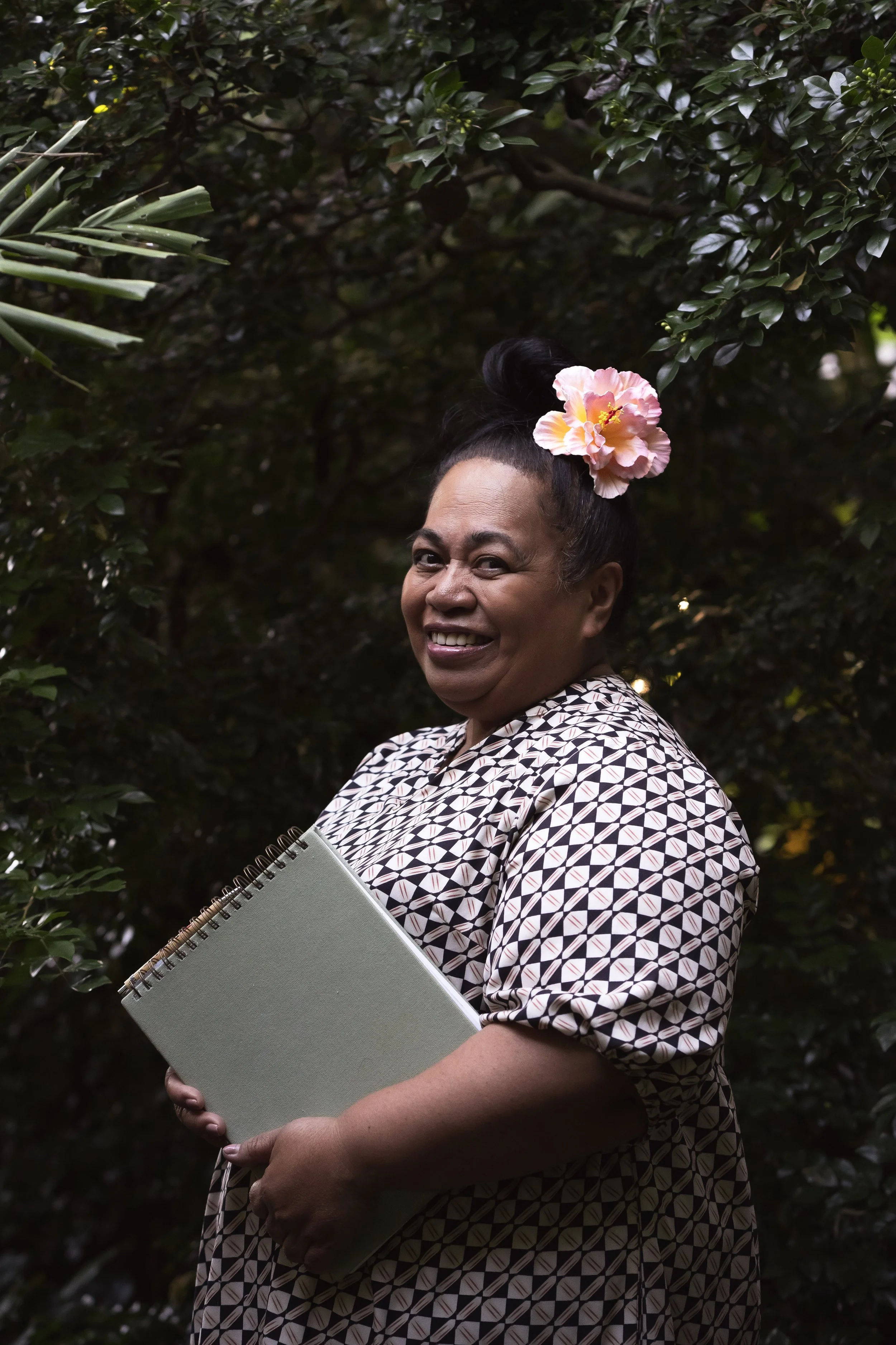 A smiling woman with dark hair styled in an updo with a pink flower accessory, holding a spiral notebook, standing outdoors in front of lush dark green foliage.