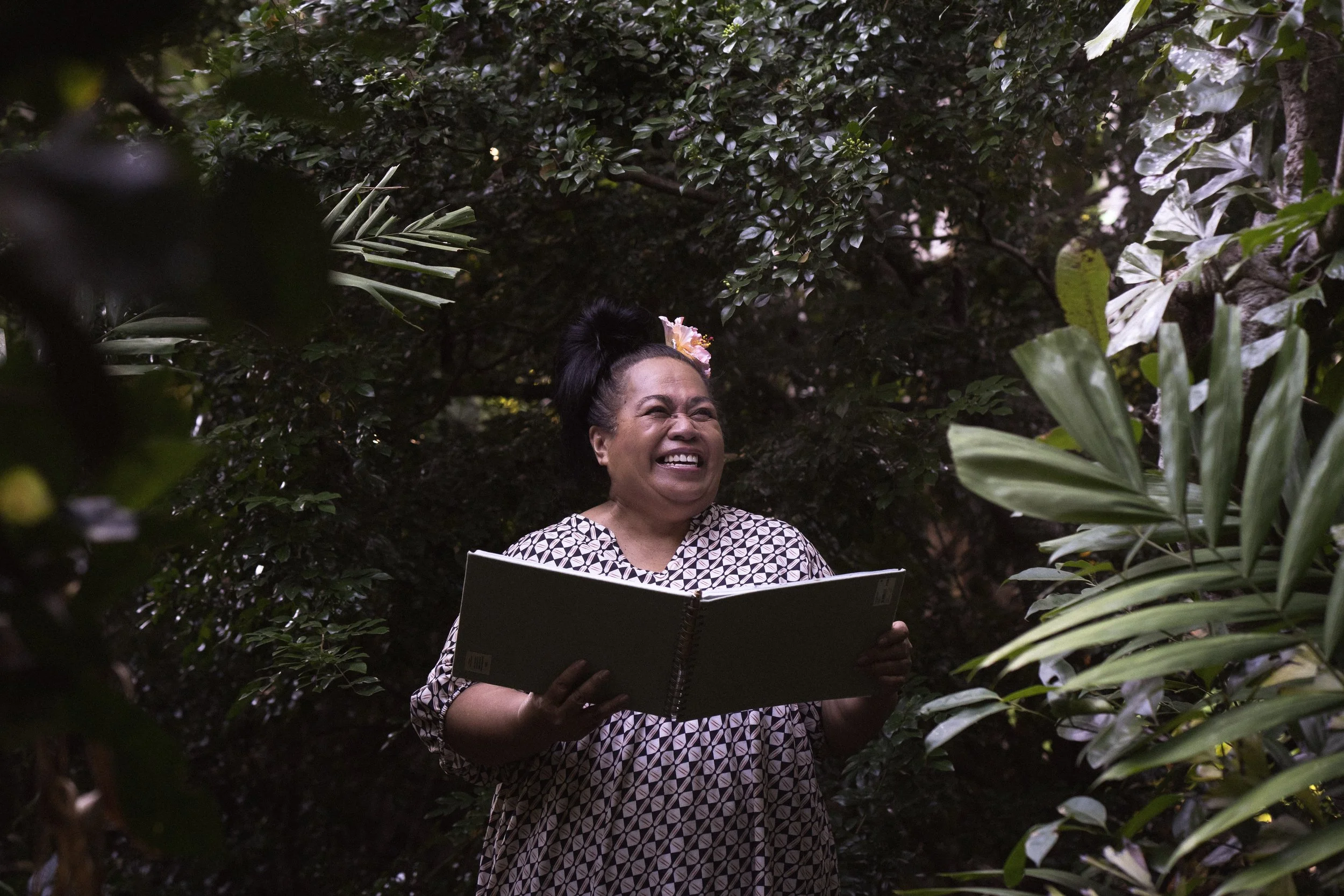 Vienna Richards smiling and laughing, holding an open notebook, surrounded by lush green foliage at home.
