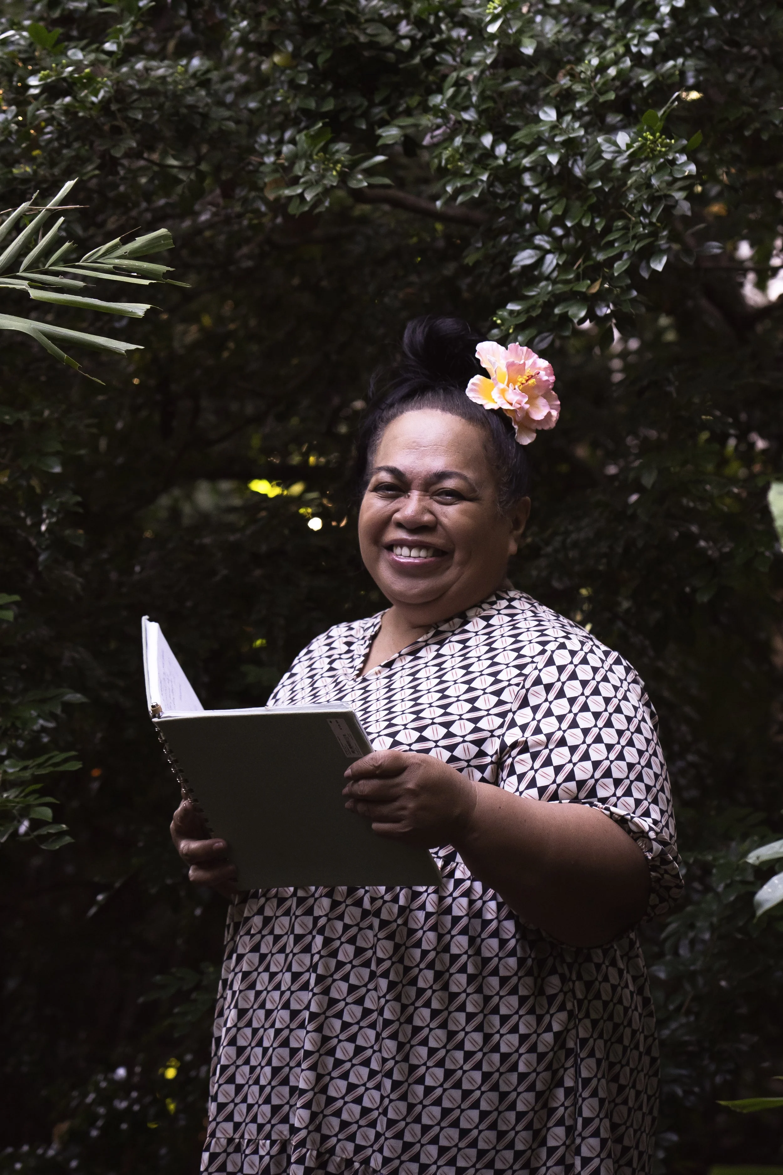 A smiling woman wearing a patterned dress and a pink flower in her hair, standing outdoors among green foliage, holding a notebook.