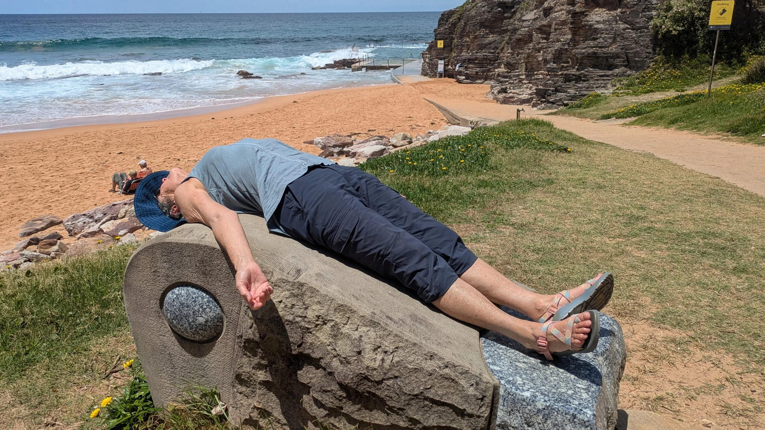 An elderly man lays on a large, rounded rock near a beach, resting with his mouth open and eyes closed. He is wearing a blue hat, a gray shirt, black shorts, and sandals. The background features a sandy beach, ocean waves, a walkway, and a sign on th