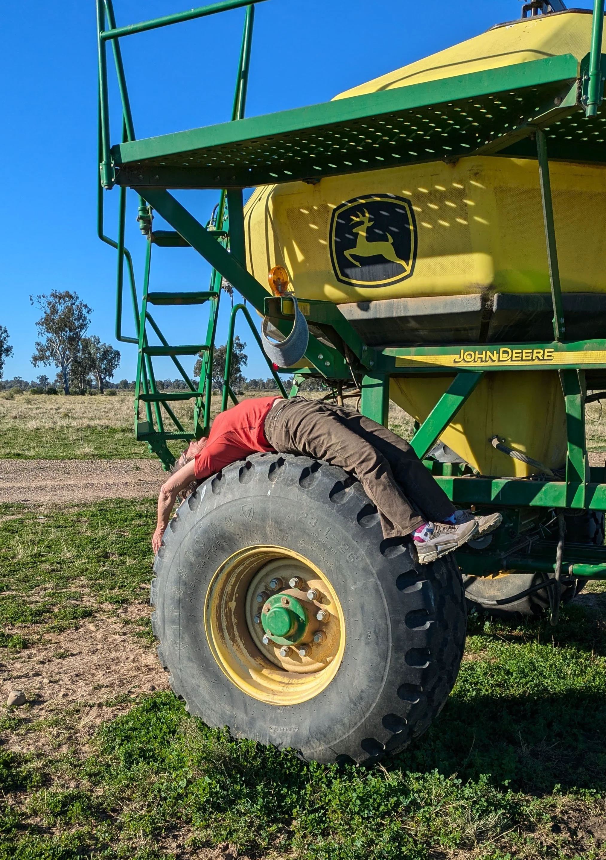 Person leaning against a large John Deere farm sprayer, head and upper body underneath the equipment, outdoors on a farm field with trees and clear blue sky.