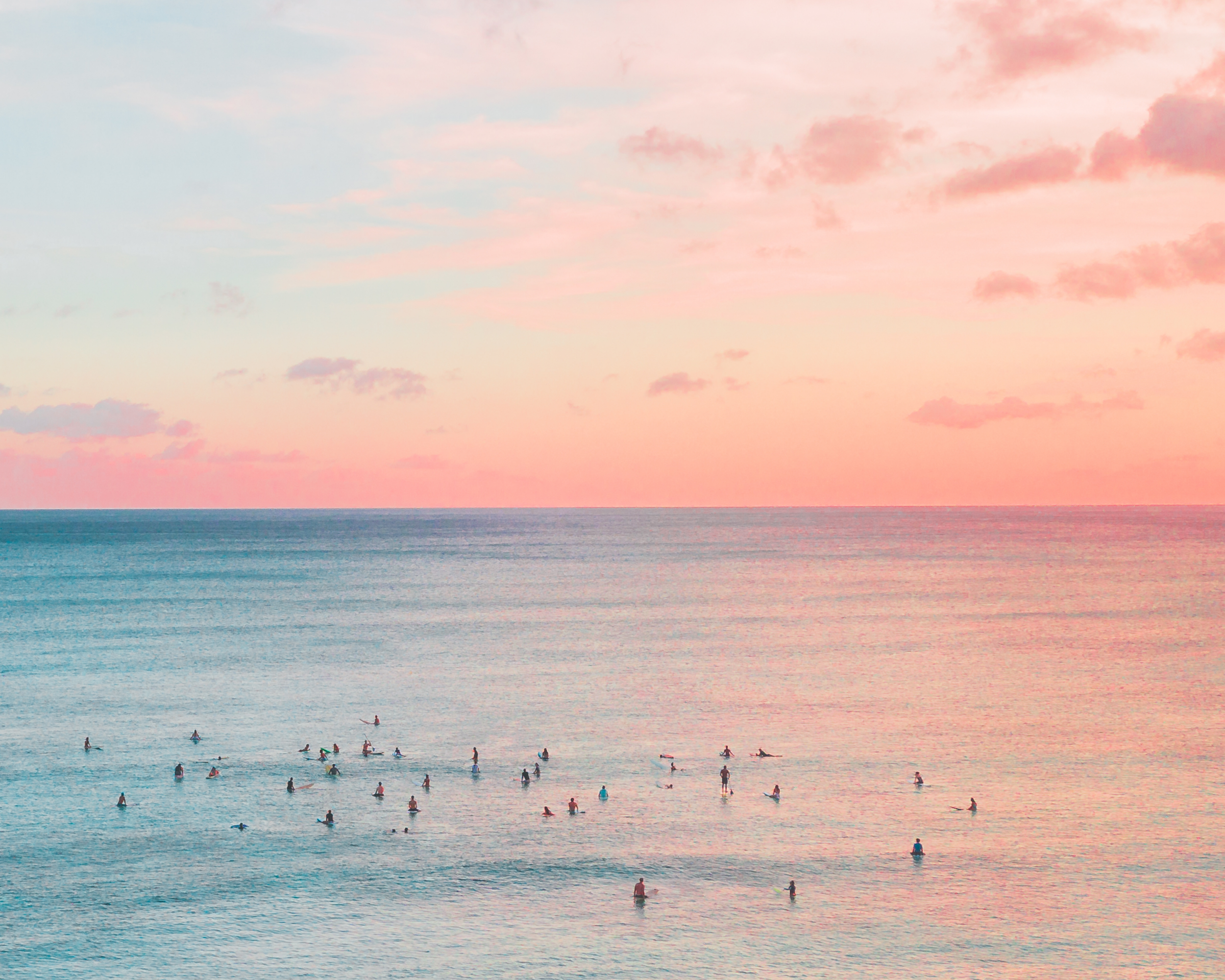Sunset over calm ocean waters with people surfing and swimming, sky with pink and orange clouds.