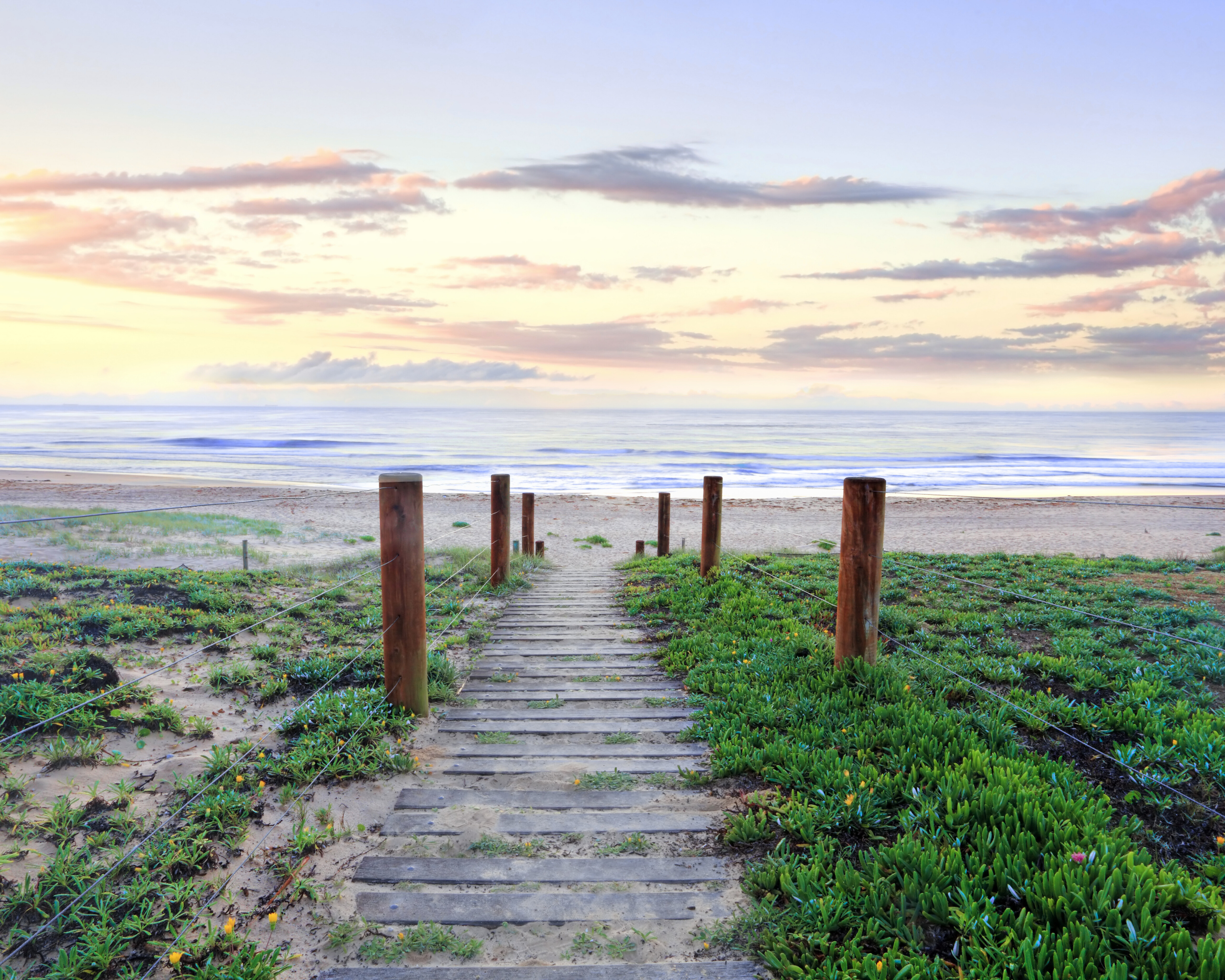 A wooden pathway leading from beachside greenery to the sandy shore, with the ocean and colorful sky at sunset in the background.