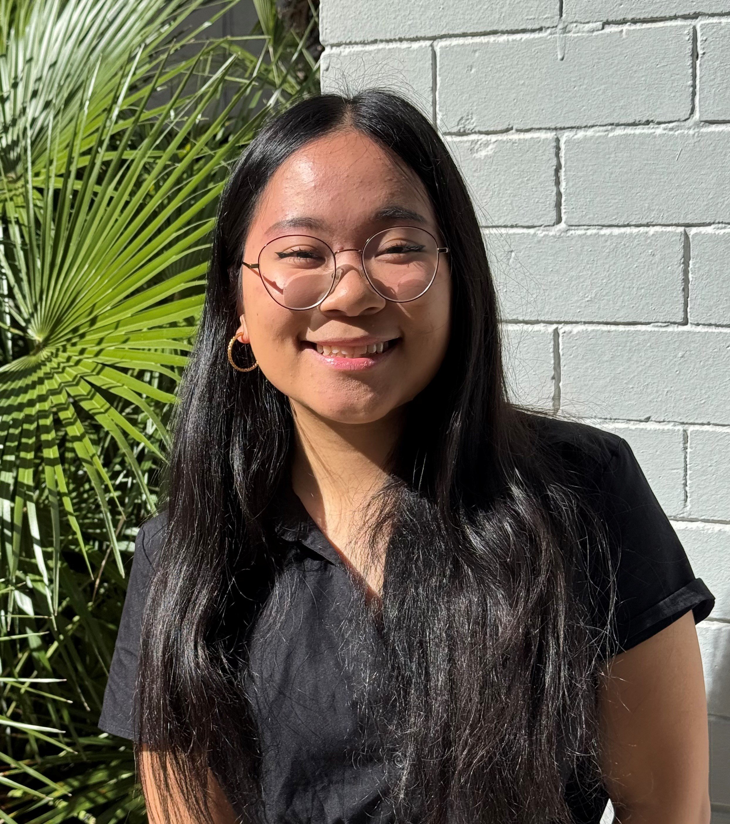 Close-up of a young woman with curly hair, wearing glasses, earrings, and layered necklaces, smiling in an indoor setting with blurred background.