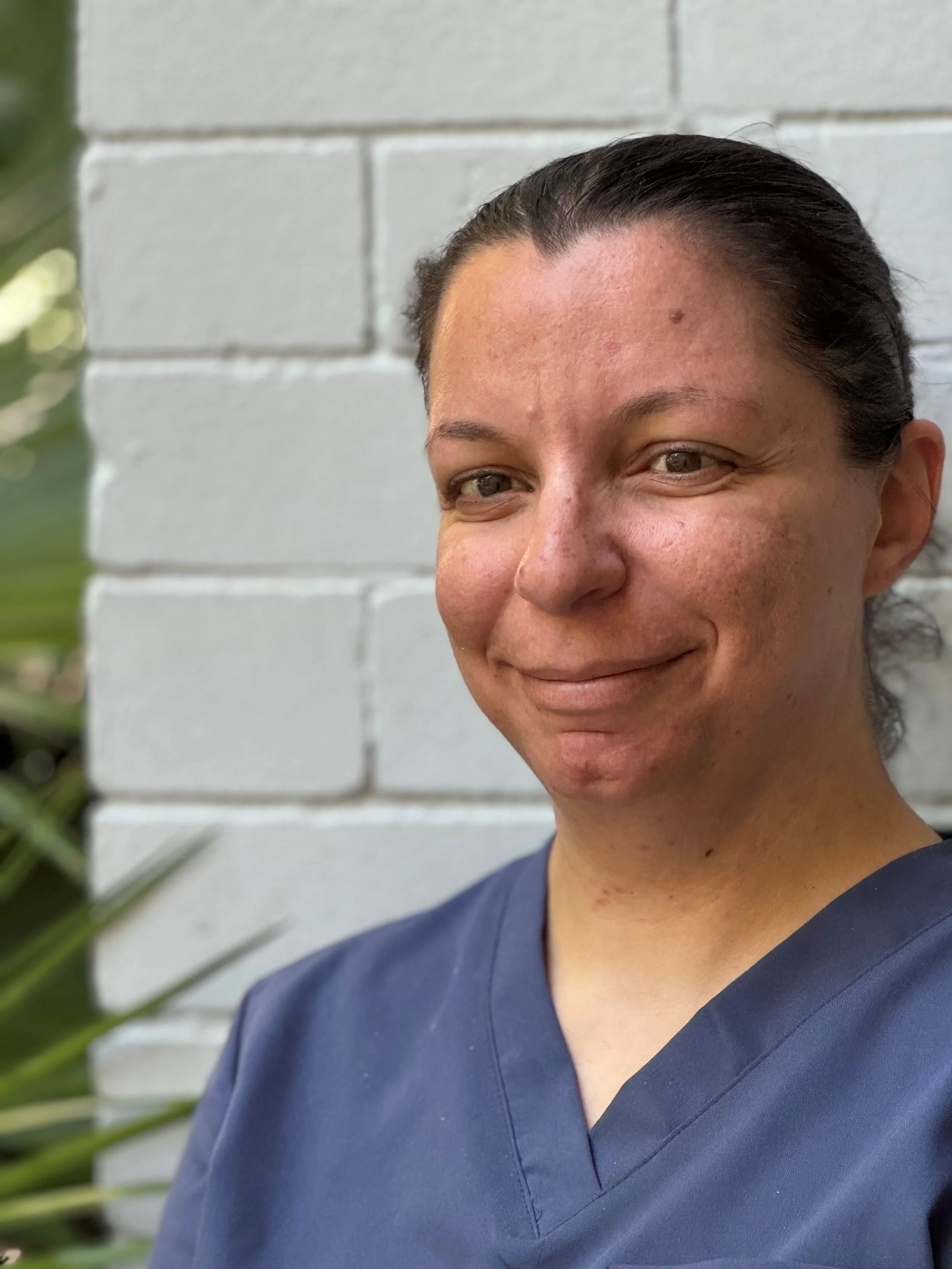 A smiling woman with dark brown curly hair, wearing a red long-sleeve turtleneck, standing outdoors with a blurred background.