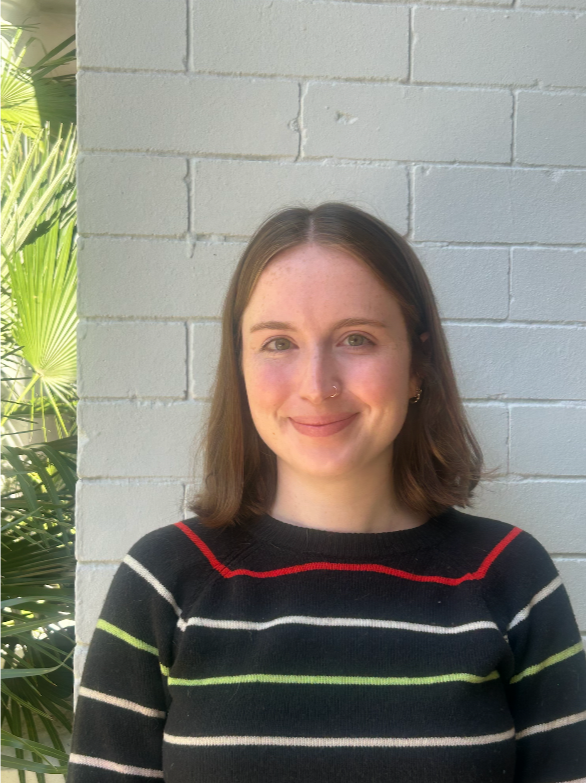 A woman with shoulder-length brown hair smiling outdoors with green foliage and a wooden fence in the background.