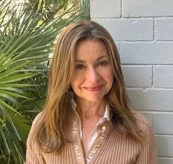 A woman with long, light brown hair, wearing a brown checkered blazer, smiling slightly, standing against a plain white wall.