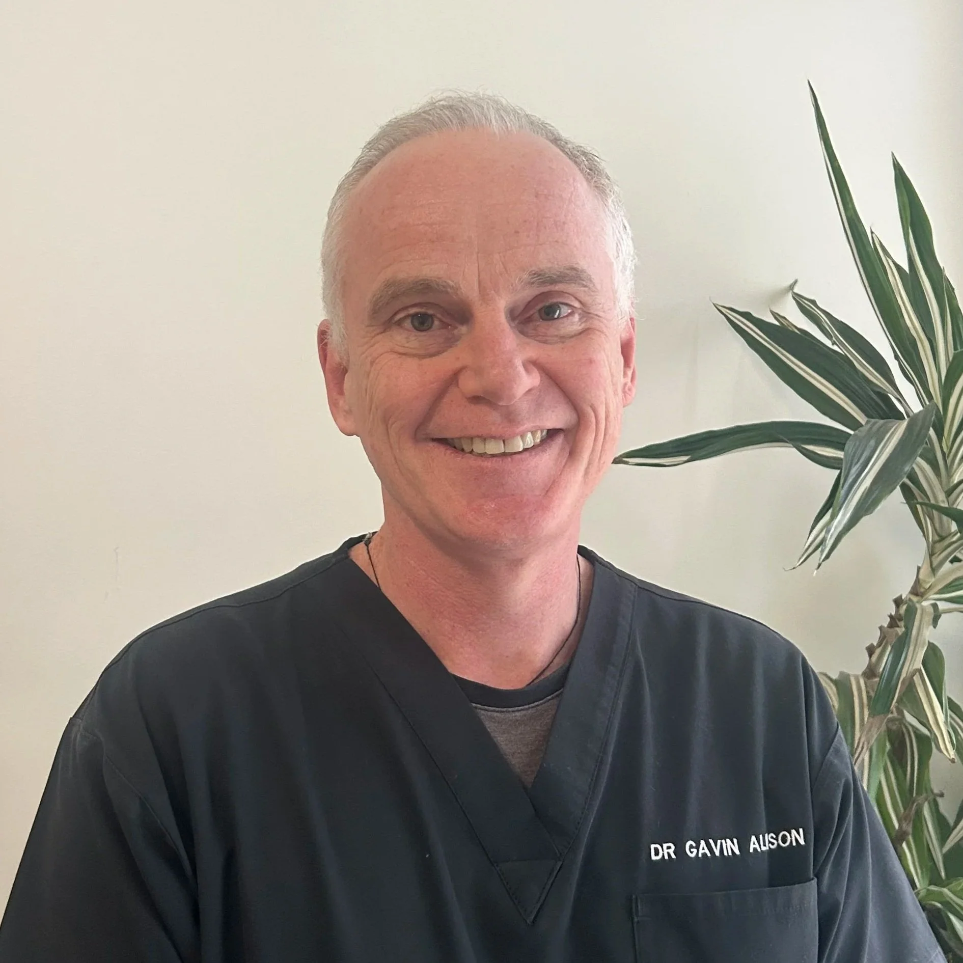 A smiling man with short gray hair wearing black medical scrubs with a name badge that reads Dr. Gavin Alison, standing in front of a plain light-colored wall with a large green leafy plant to his right.