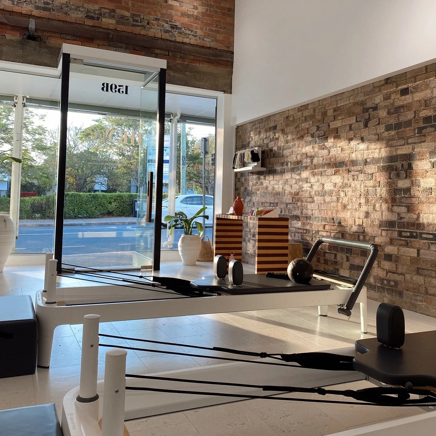 Minimalist Pilates studio with reformer machines, a brick wall, potted plants, and large glass windows letting in natural light.