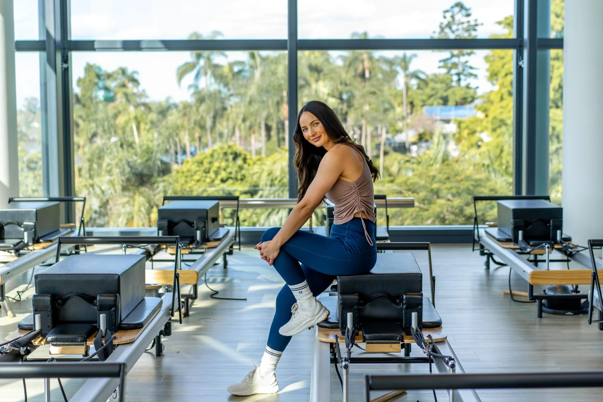 Woman sitting on a Pilates reformer in a fitness studio with large windows and greenery outside.
