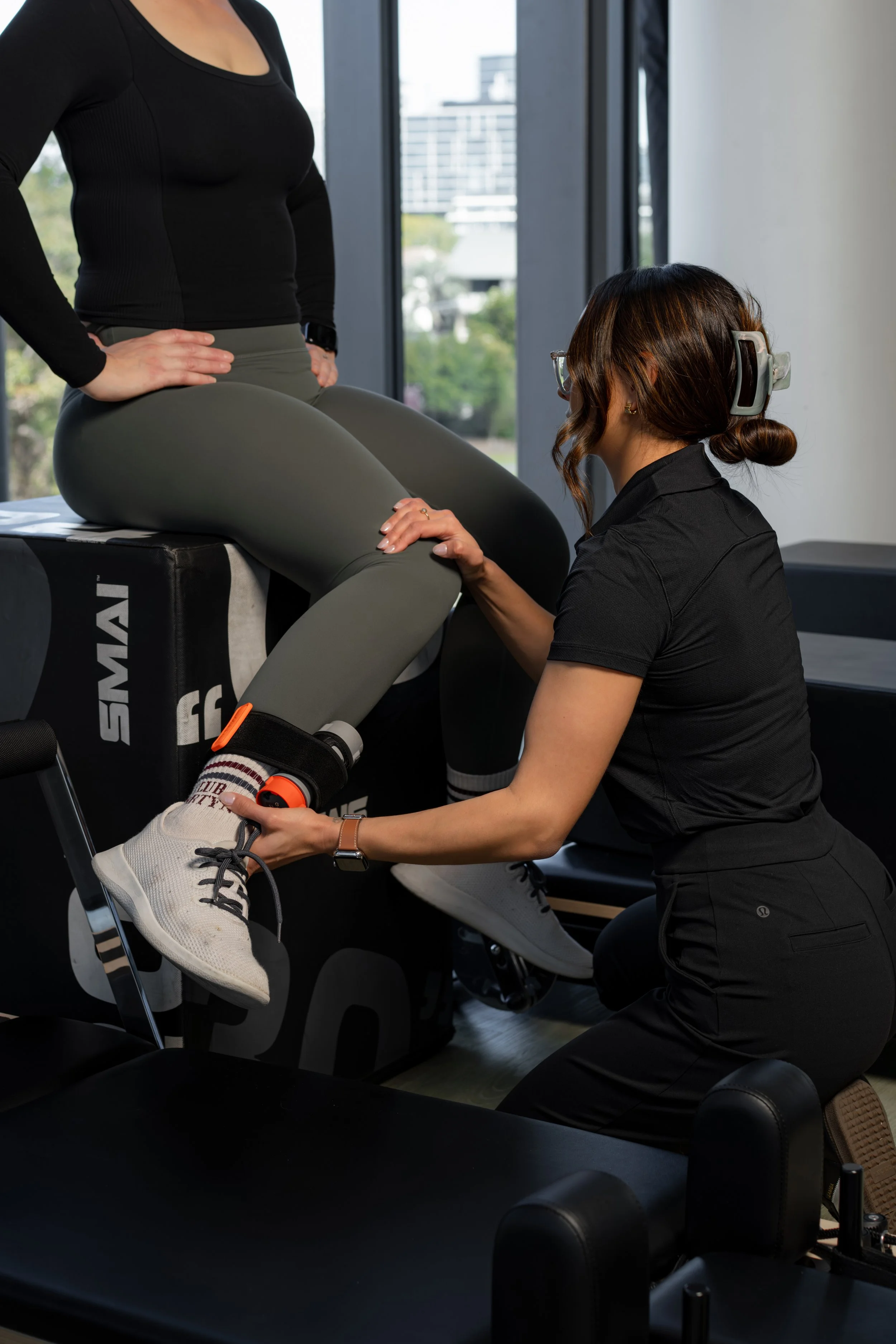 A woman in workout attire sitting on a mat, holding a circular exercise ring with both hands, in front of a rack of dumbbells in a gym.