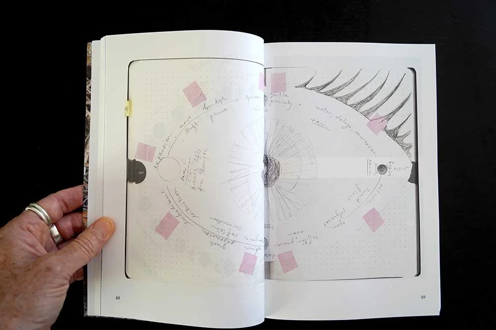 Open notebook with sketches and handwritten notes, featuring a circular diagram and a detailed eyelash sketch on the right page, held by a hand with wedding rings.