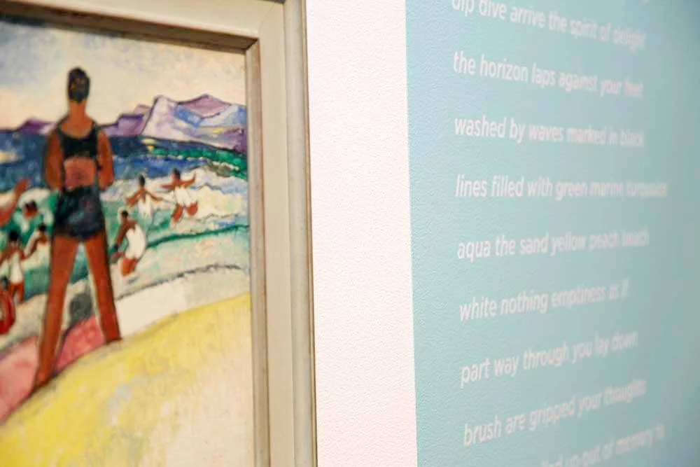 A colorful painting of a woman in a swimsuit on a beach with mountains in the background, next to a blue wall with text.