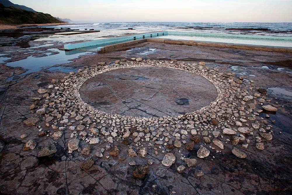 A large circular stone structure on a rocky beach, surrounded by smaller stones, with the ocean and hills in the background.