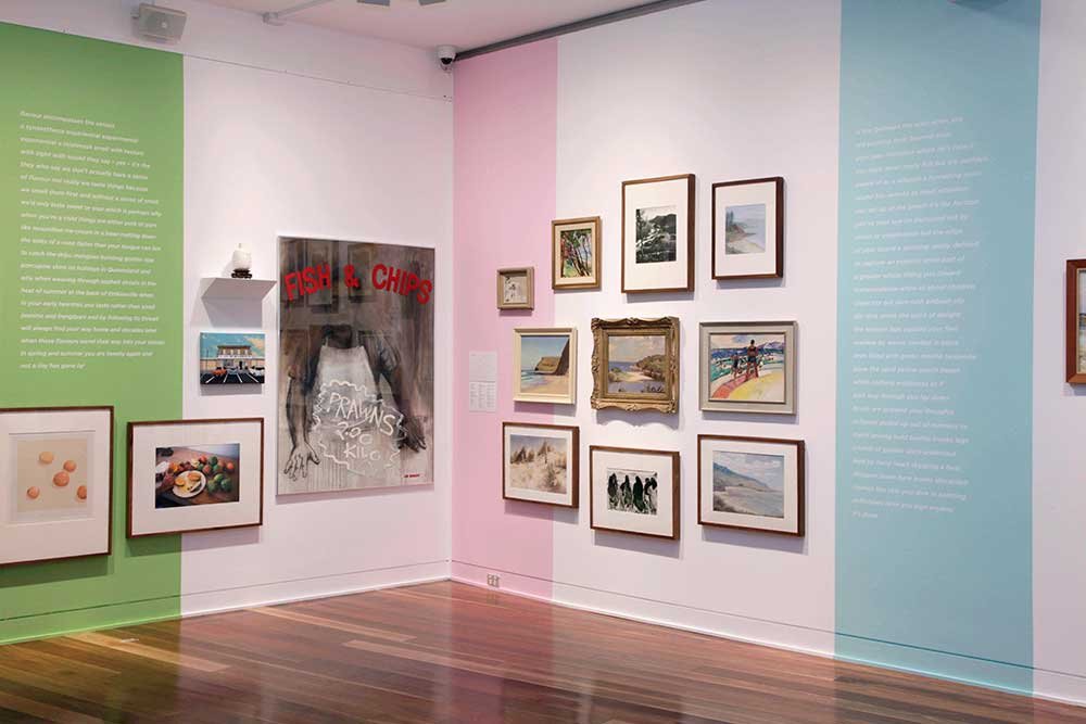 An art gallery featuring a variety of framed paintings and photographs on pastel-colored walls, with green, pink, and blue sections, and a wooden floor.