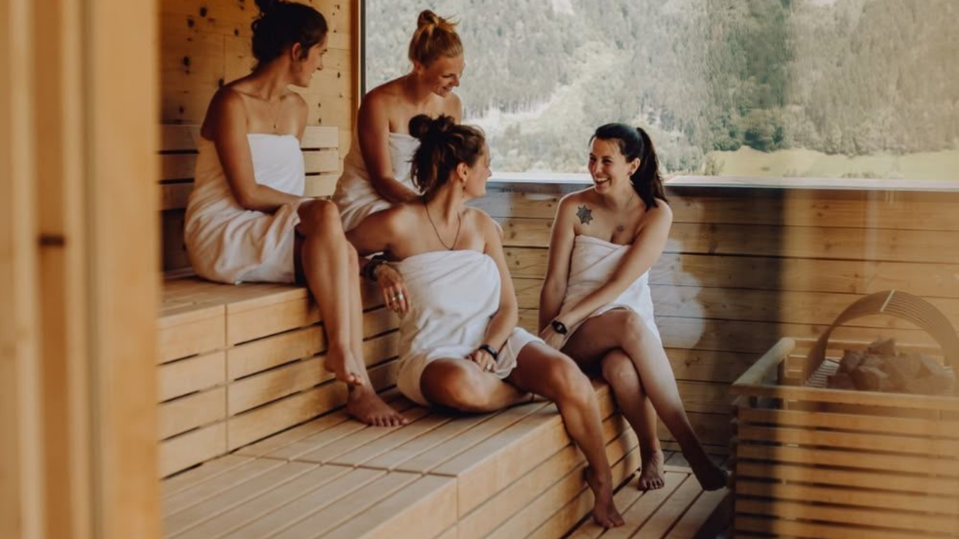 Four women in a sauna wrapped in towels, sitting and chatting happily.