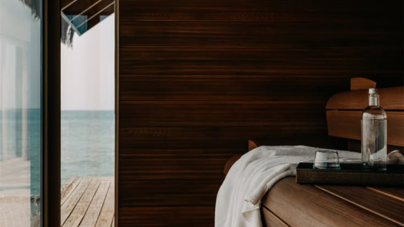 Interior of a bedroom with a view of the ocean through large sliding glass doors, featuring a wooden headboard, a glass bottle of water, and a glass on a tray on the bed.