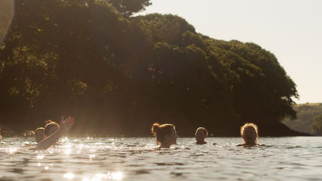 People swimming and socializing in a river during sunset with hills and trees in the background.