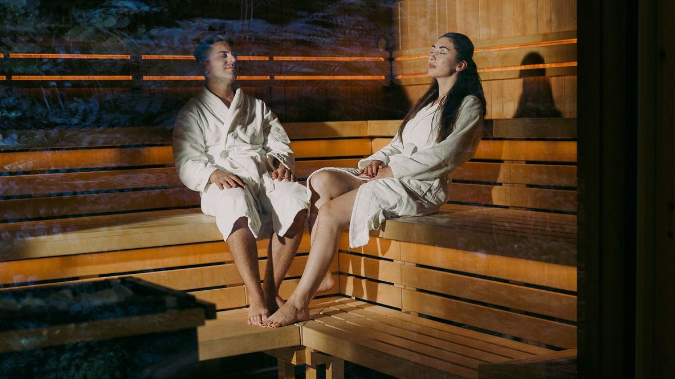 A man and a woman sitting inside a wooden sauna, wearing white robes, with the man smiling and the woman with her eyes closed.