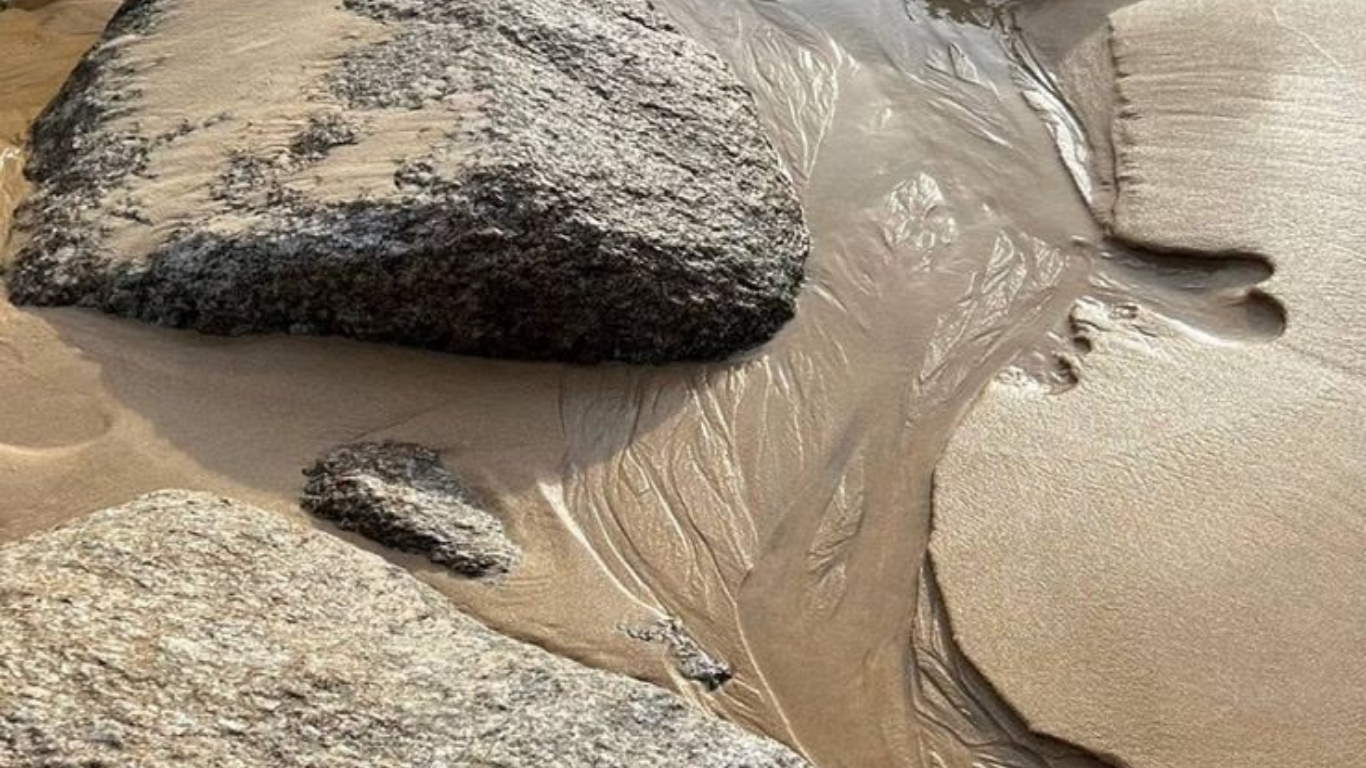 Close-up of rocks and sand on a beach, with water flowing around the rocks creating ripples.