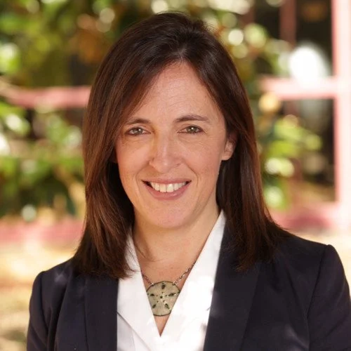 Headshot of a woman with shoulder-length brown hair, smiling, wearing a dark blazer and a white shirt, with a blurred outdoor background.